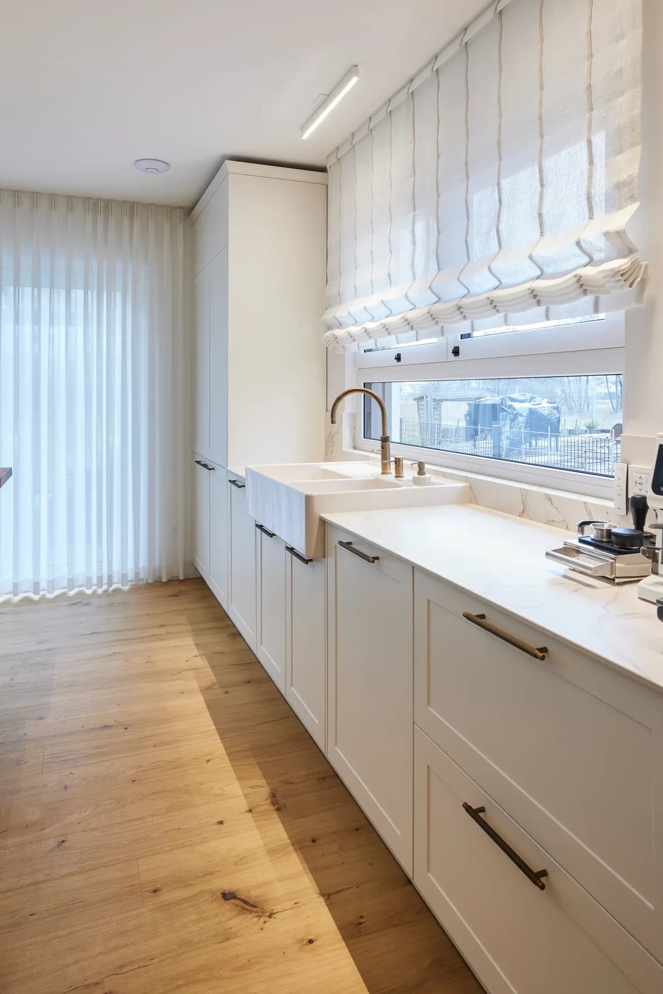 Arbeitszeile am Fensterband mit weißer Keramikspüle, Messing-Armatur und cremeweißen Rahmenfronten, davor helle Vorhänge und Eichendielen.