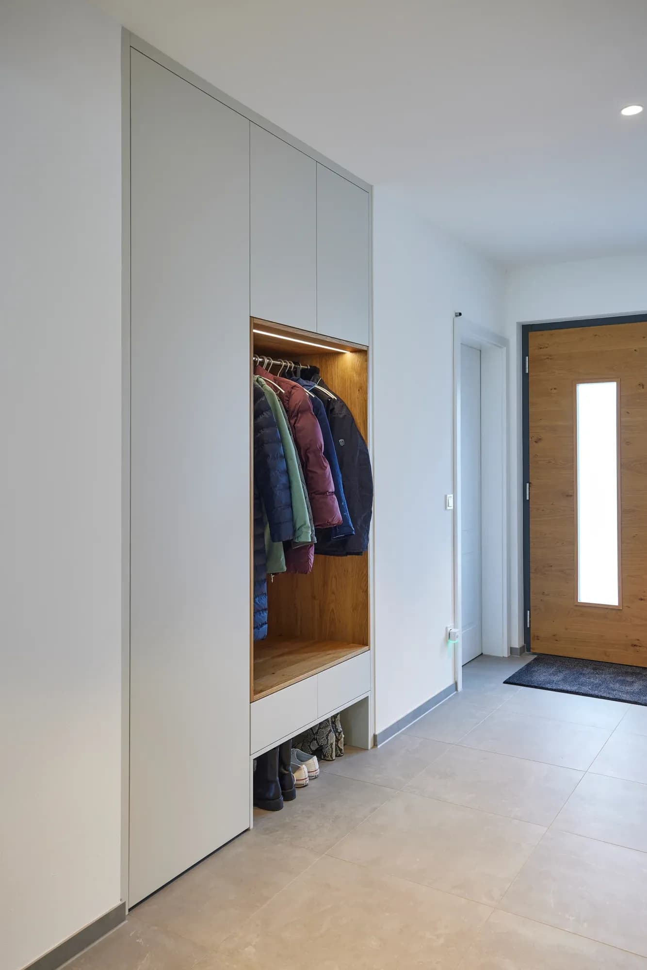 Eingebaute Garderobe im Eingang: helle Fronten mit eichengefütterter, beleuchteter Nische, Sitzbank und Schuhauszug, daneben die Haustür.