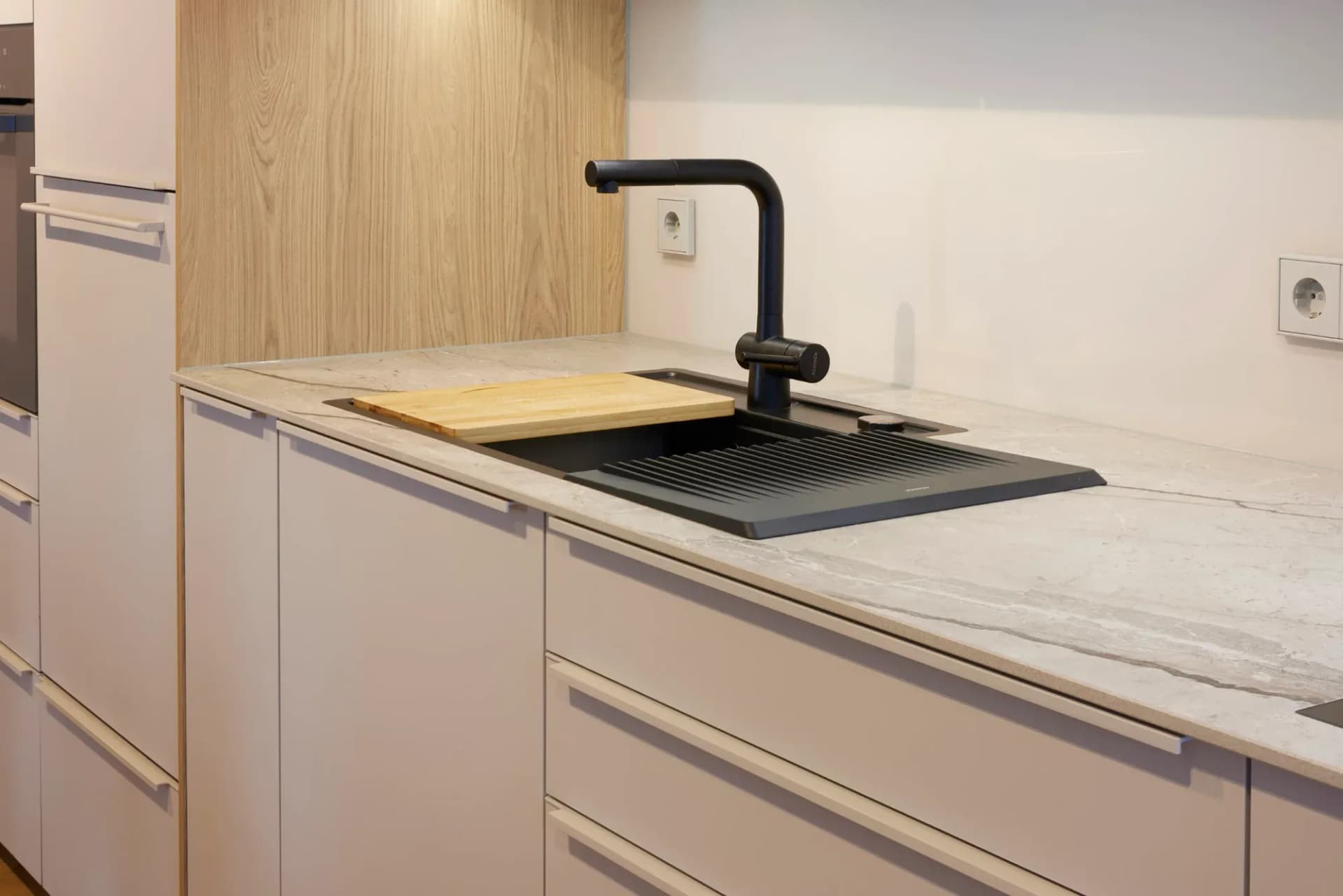 Detail der Arbeitsfläche: heller Naturstein mit feiner Aderung, schwarze Schock-Spüle mit Holzschneidbrett und schwarze Armatur vor Asteiche-Rückwand.