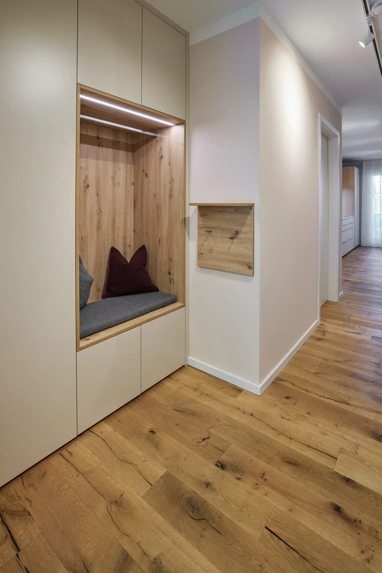 Eingebaute Garderobe im Flur: beleuchtete Nische mit Sitzbank und Kissen, Kleiderstange und Asteiche-Rückwand, dazu ein Hakenbrett aus Eiche.