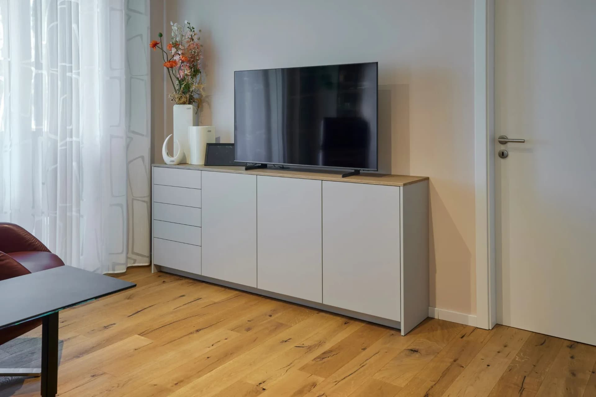 Sideboard im Wohnbereich mit Asteiche-Deckplatte und sandfarbenen Fronten, darauf ein Fernseher und Trockenblumen in weißen Vasen.