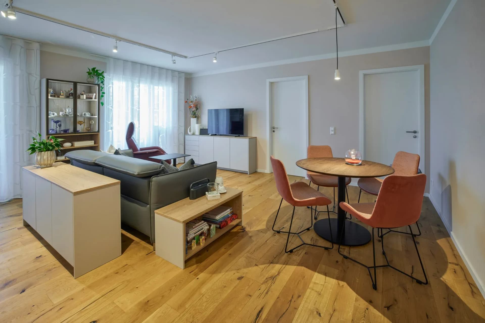 Offener Wohn- und Essbereich: Sideboard mit Fernseher, Glasvitrine, Sofa und runder Esstisch mit terrakottafarbenen Stühlen auf Eichendielenboden.