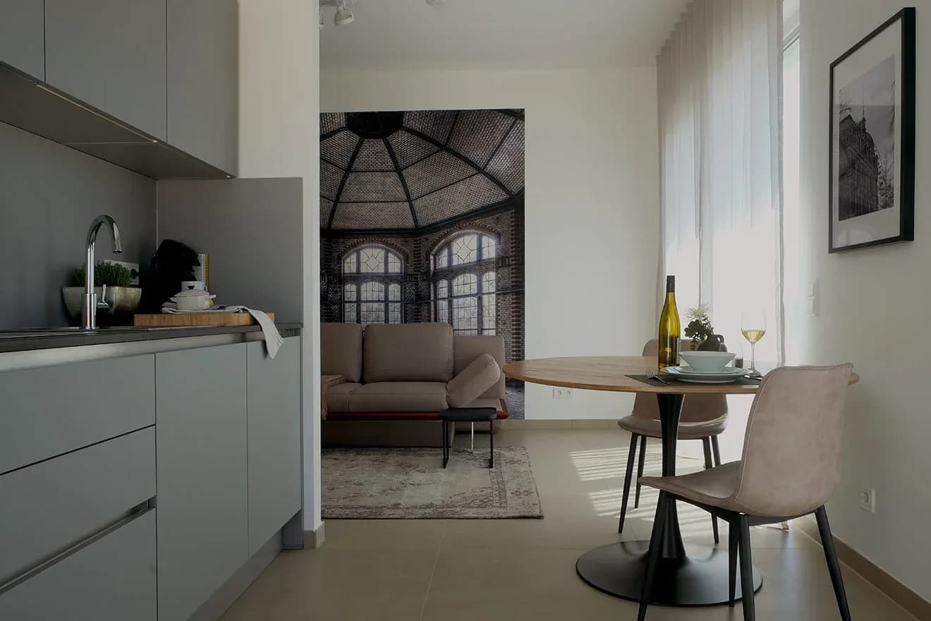 Serviced-Apartment im Reuter Quartier 33 in Landau: kompakte Kochnische mit grifflosen grauen Fronten, runder Essplatz mit zwei Stühlen und großformatiges Wandbild eines Glasdach-Pavillons.