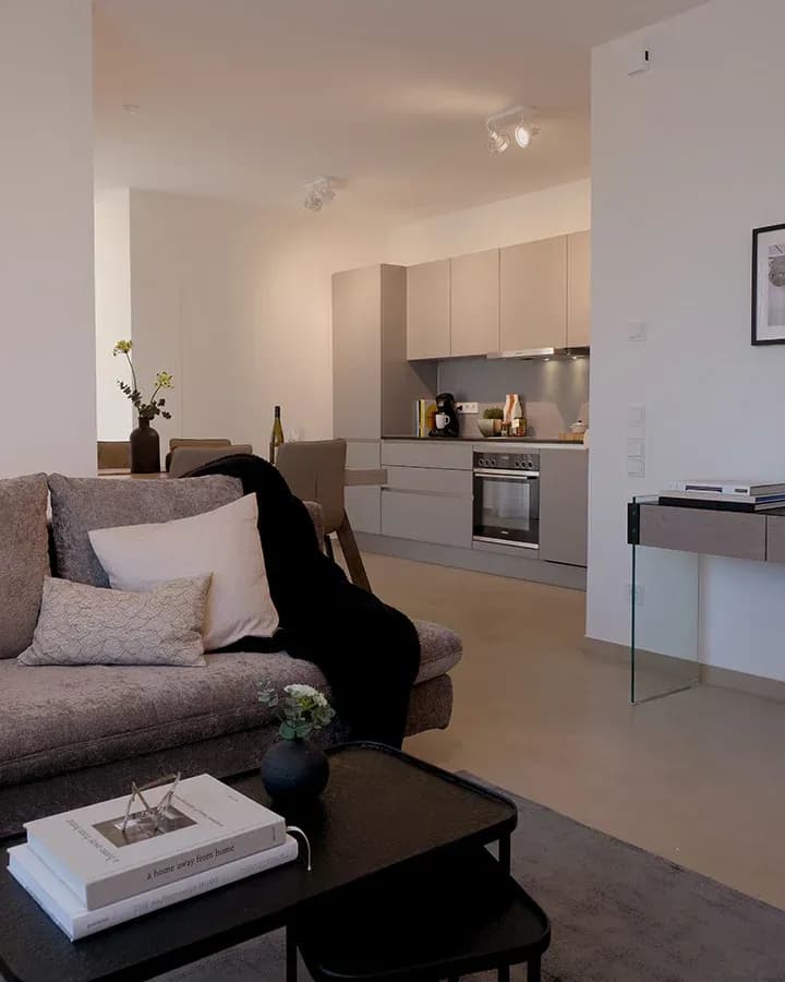 Offene Wohnküche eines Serviced-Apartments am Abend: Sofa mit Kissen im Vordergrund, grifflose Küchenzeile mit Einbauherd im Hintergrund.