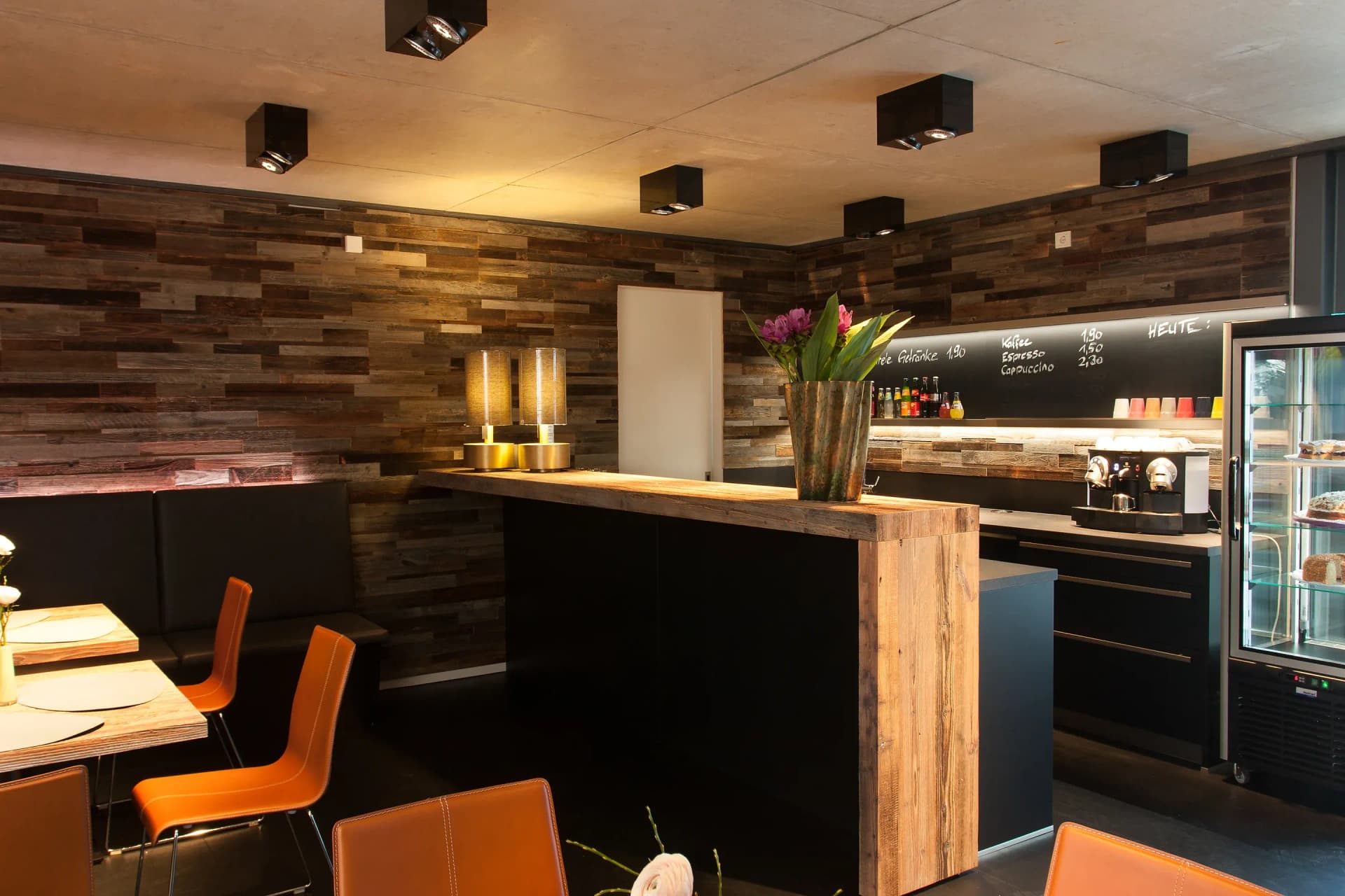 Café-Ecke eines Ladengeschäfts: freistehende Theke mit schwarzem Korpus und massiver Altholz-Auflage, dahinter Tafelwand mit Getränkekarte, Espressomaschine und gläserne Kühlvitrine; links eine Lederbank mit cognacfarbenen Stühlen unter einer Altholz-Wand.