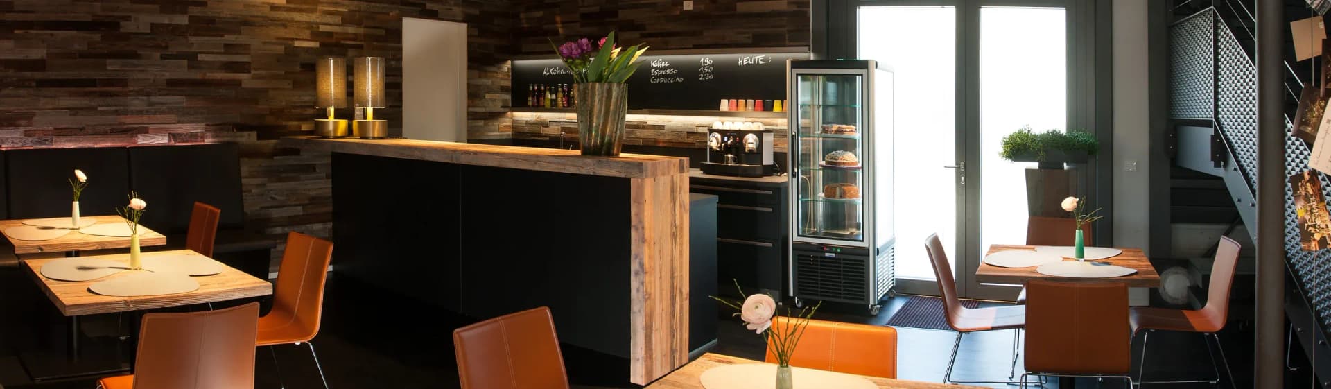 Panorama der Café-Theke: links die Sitzecke mit Tischen und Lederstühlen, mittig die freistehende Altholz-Theke, rechts die Arbeitszeile mit Kühlvitrine und die Eingangstür mit Tageslicht.