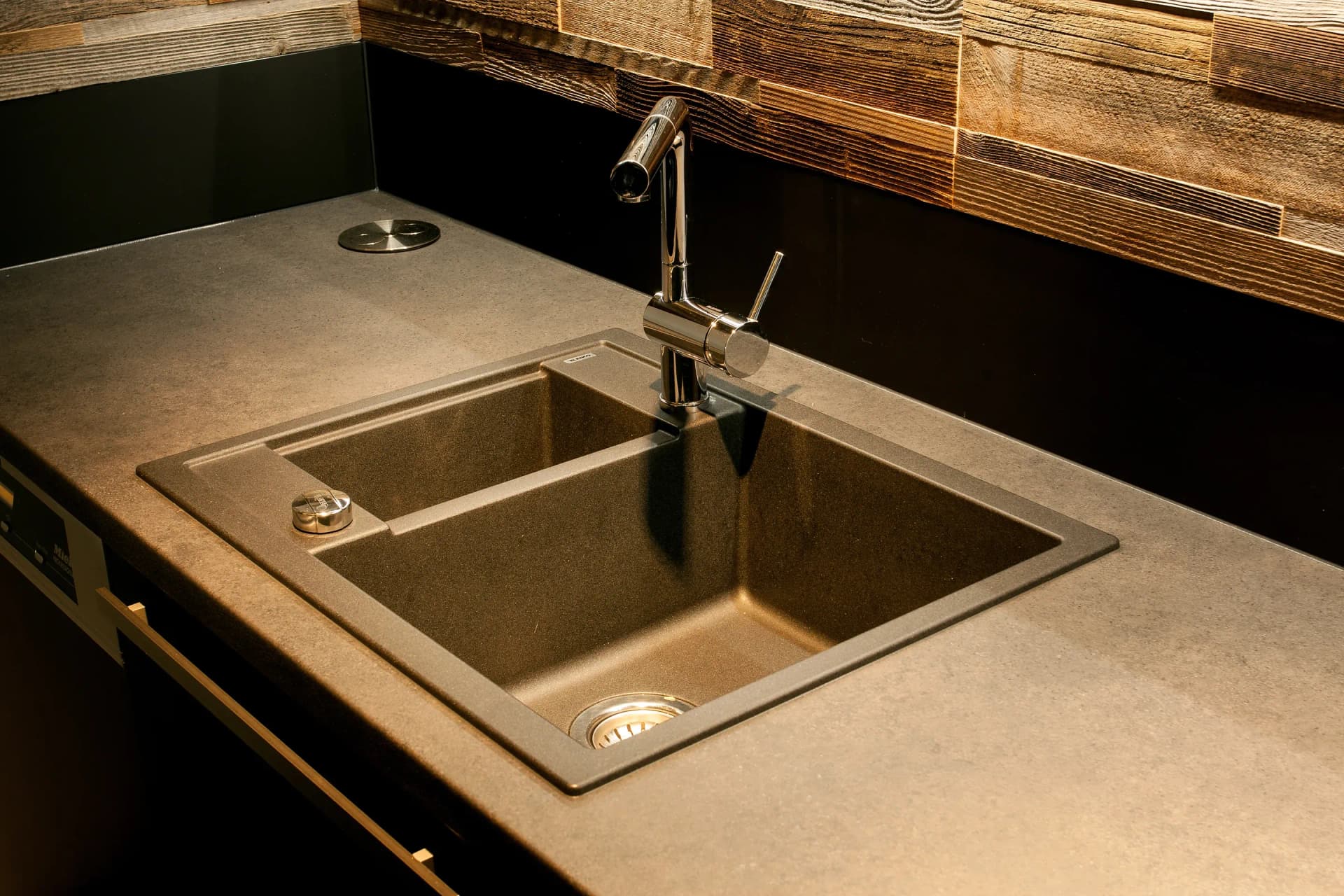 Detail der Arbeitsfläche: anthrazitfarbene Verbundstein-Spüle mit ausziehbarer Chromarmatur in einer Arbeitsplatte in Steinoptik vor Altholz-Rückwand.