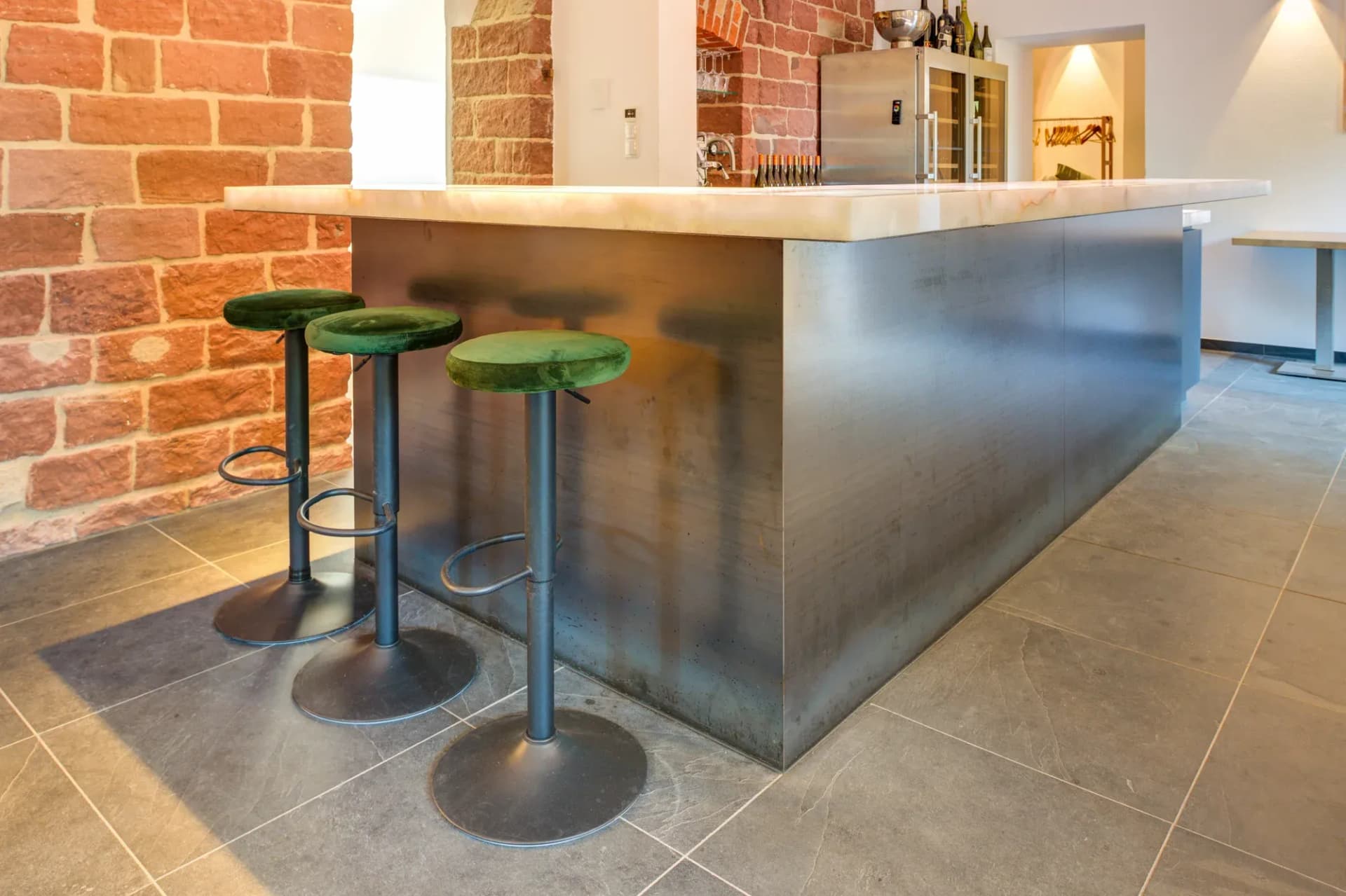Bartheke mit hinterleuchteter Onyx-Deckplatte und metallverkleideter Front, davor drei grüne Barhocker vor einer Sichtbacksteinwand.
