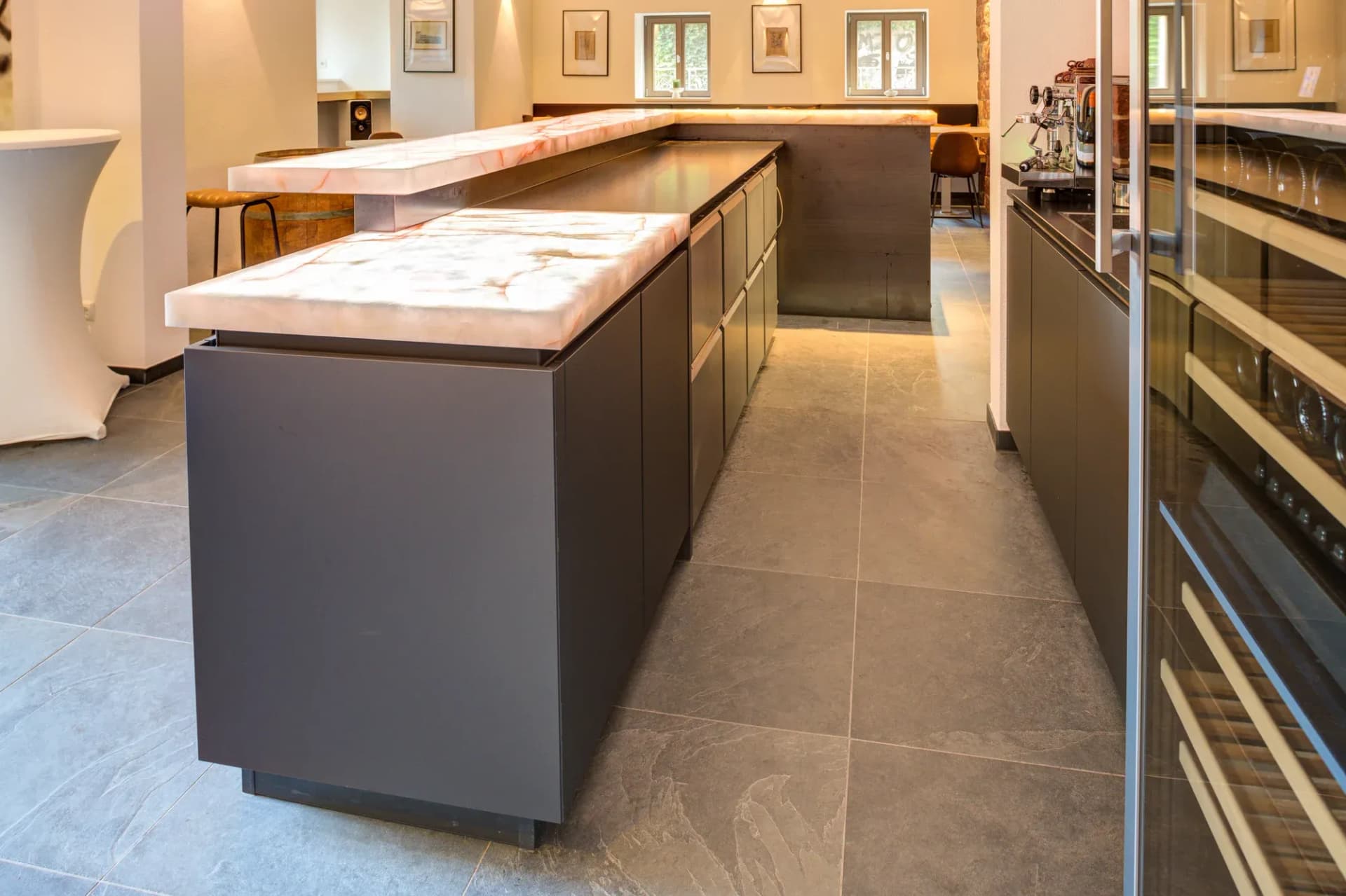 Zwei Thekenblöcke mit Onyx-Deckplatten in anthrazitfarbener Ausführung, rechts ein Weinklimaschrank, im Hintergrund Sitzplätze.