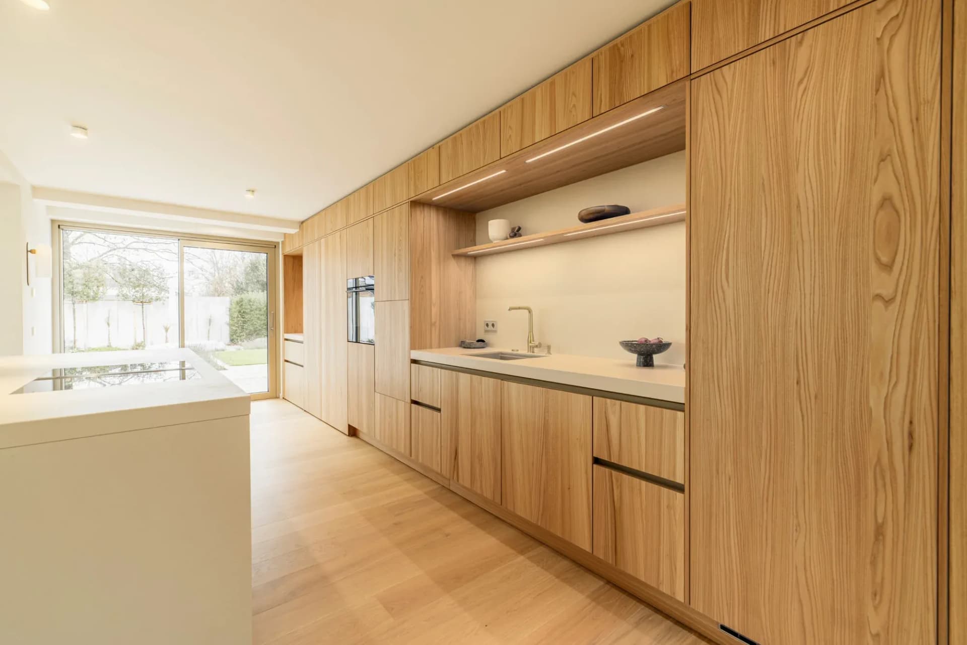 Arbeitszeile aus Holzfronten mit heller Natursteinfläche, offenem Regal mit indirekter Beleuchtung, Messingarmatur und Spüle, links die Insel.