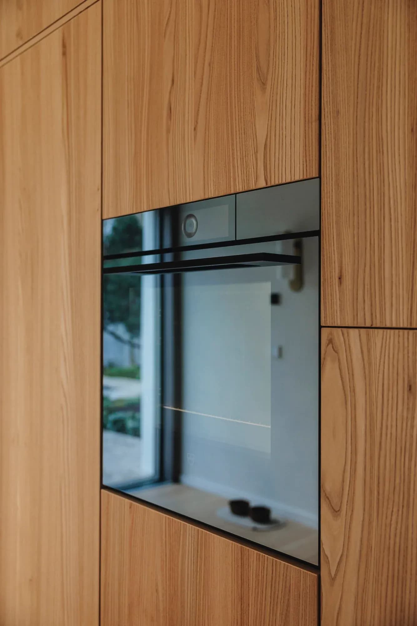 Flächenbündig in die Holzwand eingebauter Backofen mit dunkler Glasfront, in der sich die Fensterfront spiegelt.