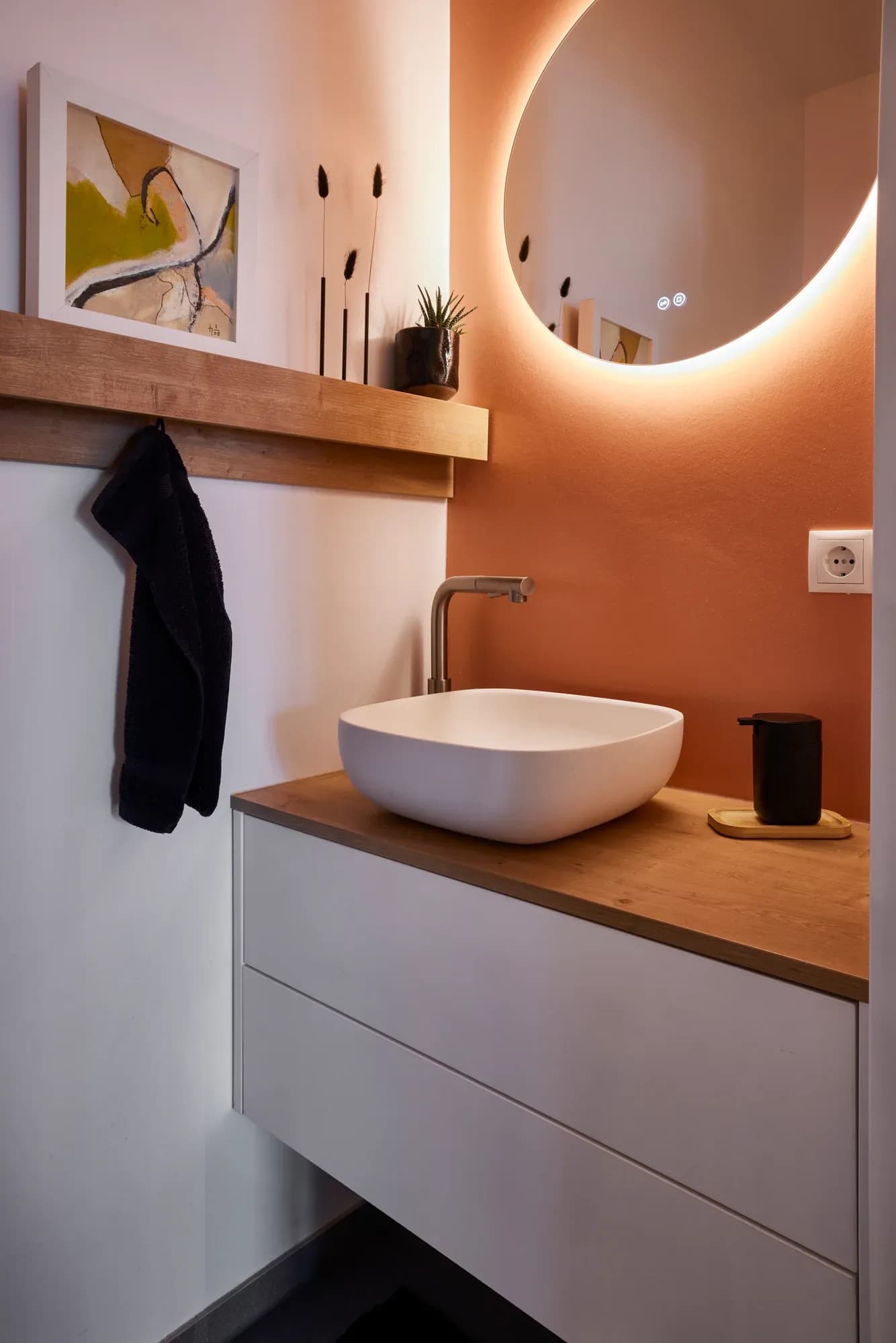Gäste-WC mit Eichen-Wandbord, Aufsatzwaschbecken auf weißem Unterschrank und rundem, hinterleuchtetem Spiegel vor terrakottafarbener Wand.