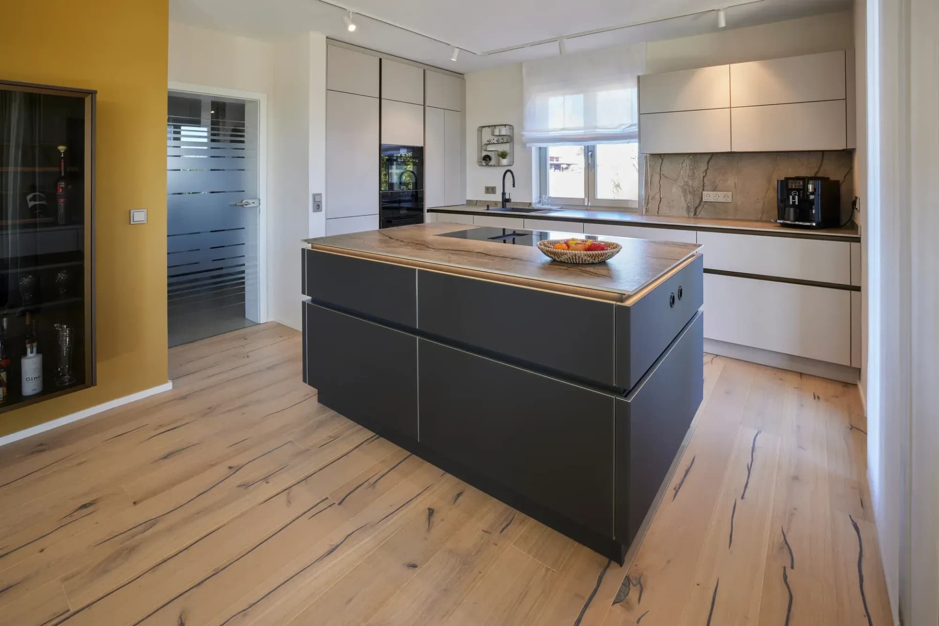 Kücheninsel mit anthrazitfarbenen Fronten und massiver Eichen-Arbeitsplatte, dahinter die helle Küchenzeile und durchgehende Eiche-Landhausdiele.