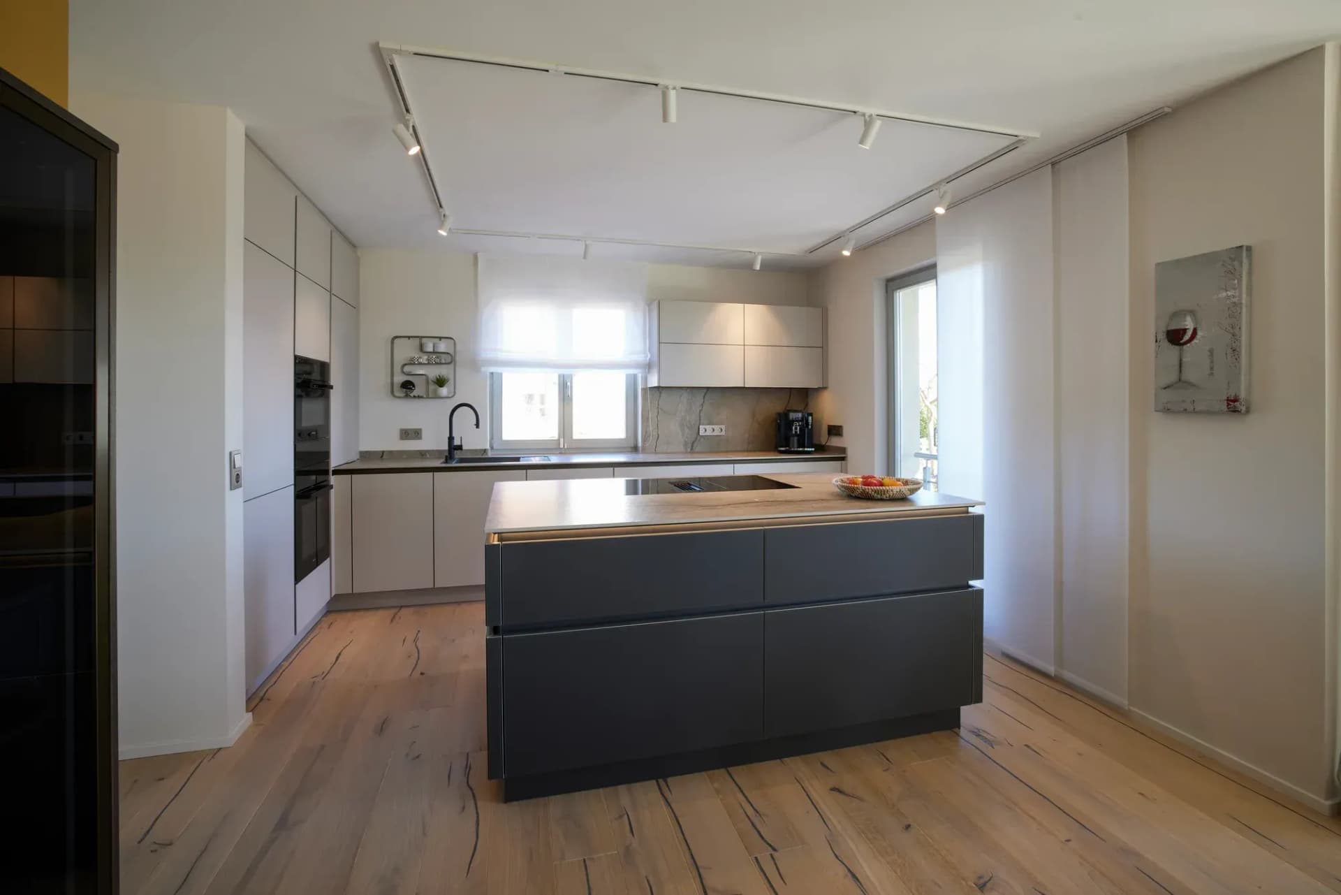 Blick durch die offene Küche: freistehende Insel in Anthrazit, helle Hochschränke und Küchenzeile am Fenster über Eiche-Landhausdiele.