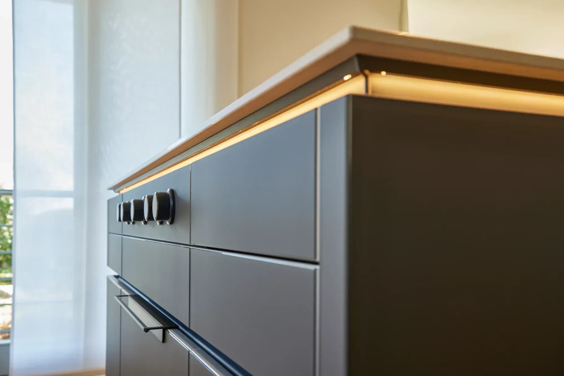 Detail der Kücheninsel: anthrazitfarbene grifflose Fronten, massive Eichenauflage mit warmer LED-Lichtfuge und die Bedienknöpfe des Kochfelds.