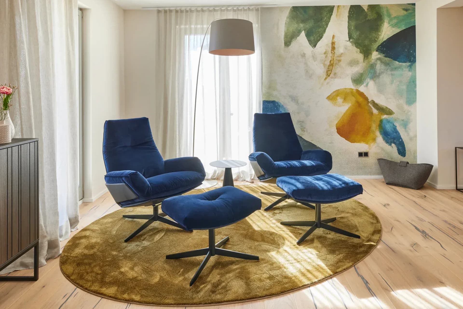 Wohnbereich mit zwei blauen Samt-Loungesesseln auf goldfarbenem Rundteppich vor einem großformatigen Aquarell-Wandbild.