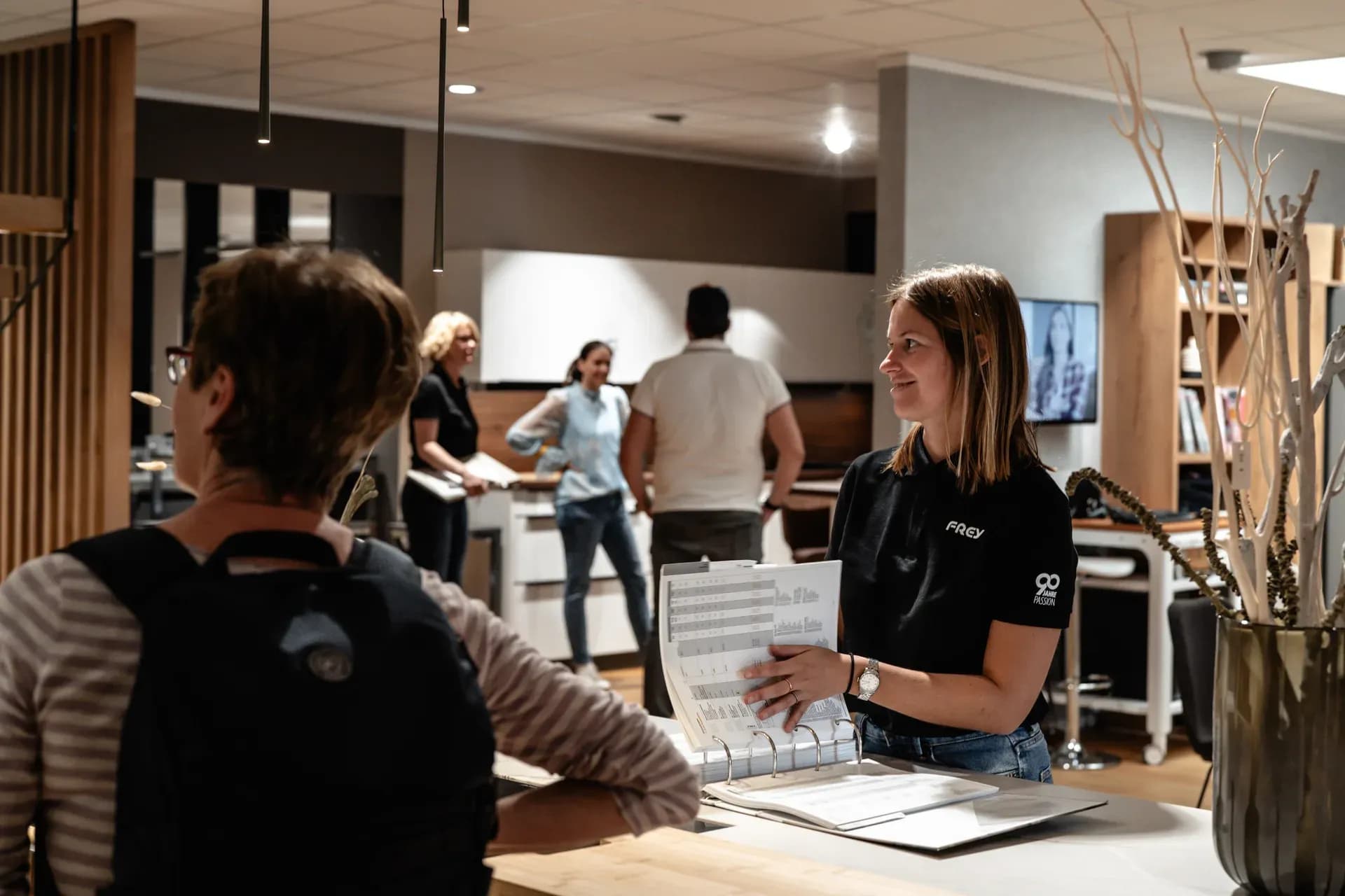 Frey-Beraterin zeigt einer Besucherin Unterlagen im Ordner, dahinter Besucher zwischen den Küchen der Ausstellung in Kandel