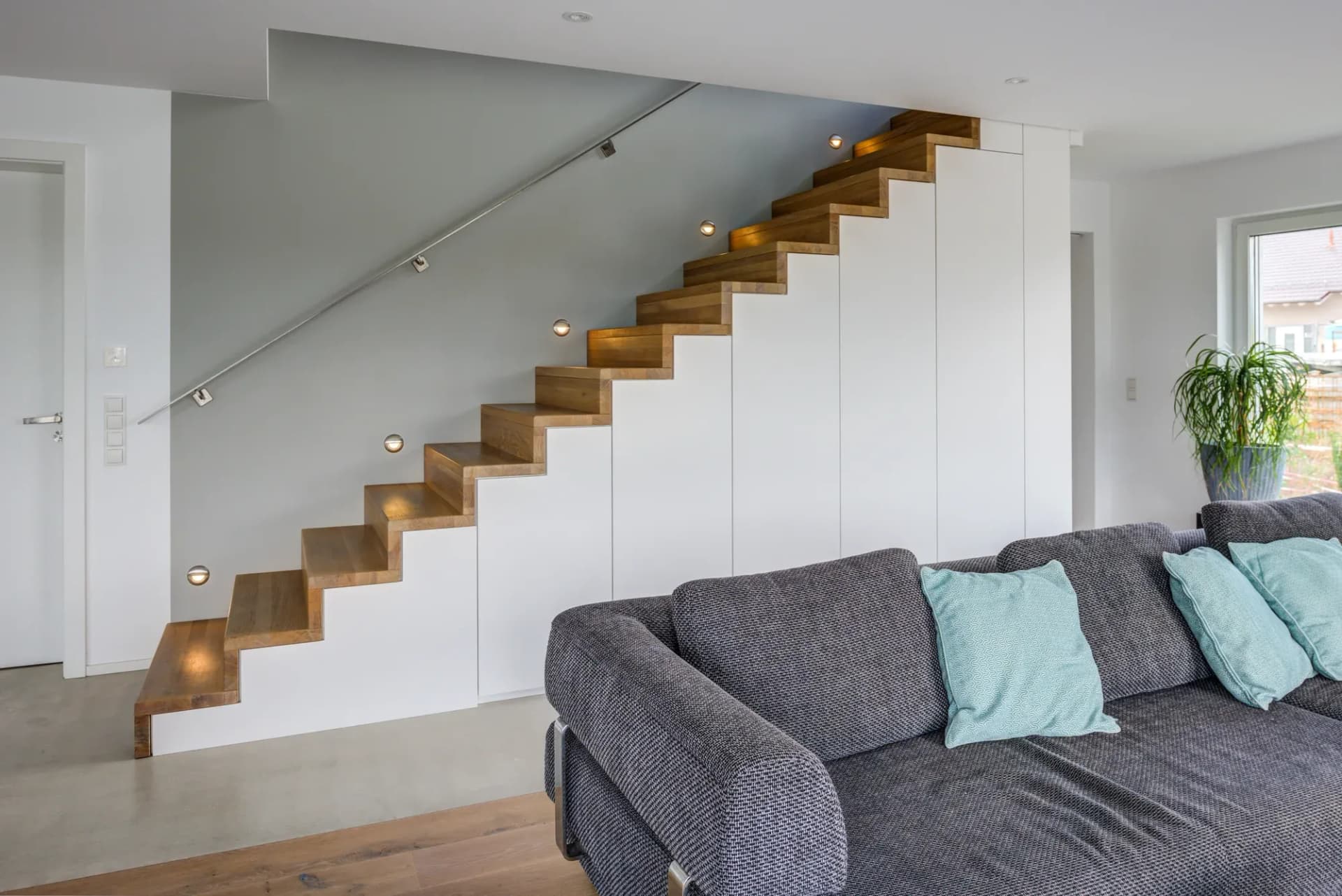 Offener Wohnraum mit einer Faltwerktreppe aus Eiche, die über einer geschlossenen weißen Schrankwand aufsteigt, dazu eingelassene Wandleuchten und ein graues Sofa.