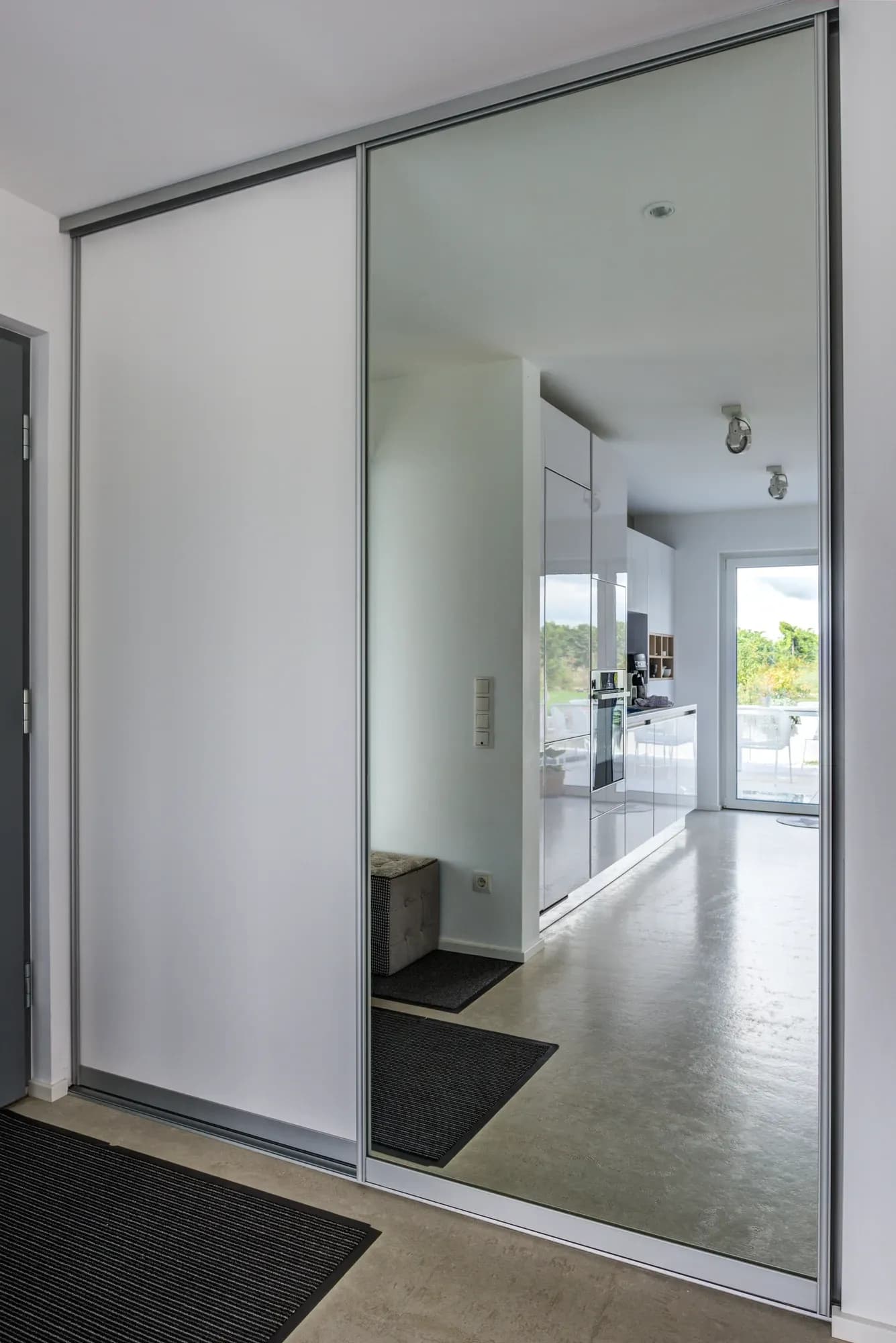 Spiegel-Schiebetür des Flurschranks, in der sich die offene weiße Küche mit Einbaubackofen und der Ausgang zur Terrasse spiegeln.