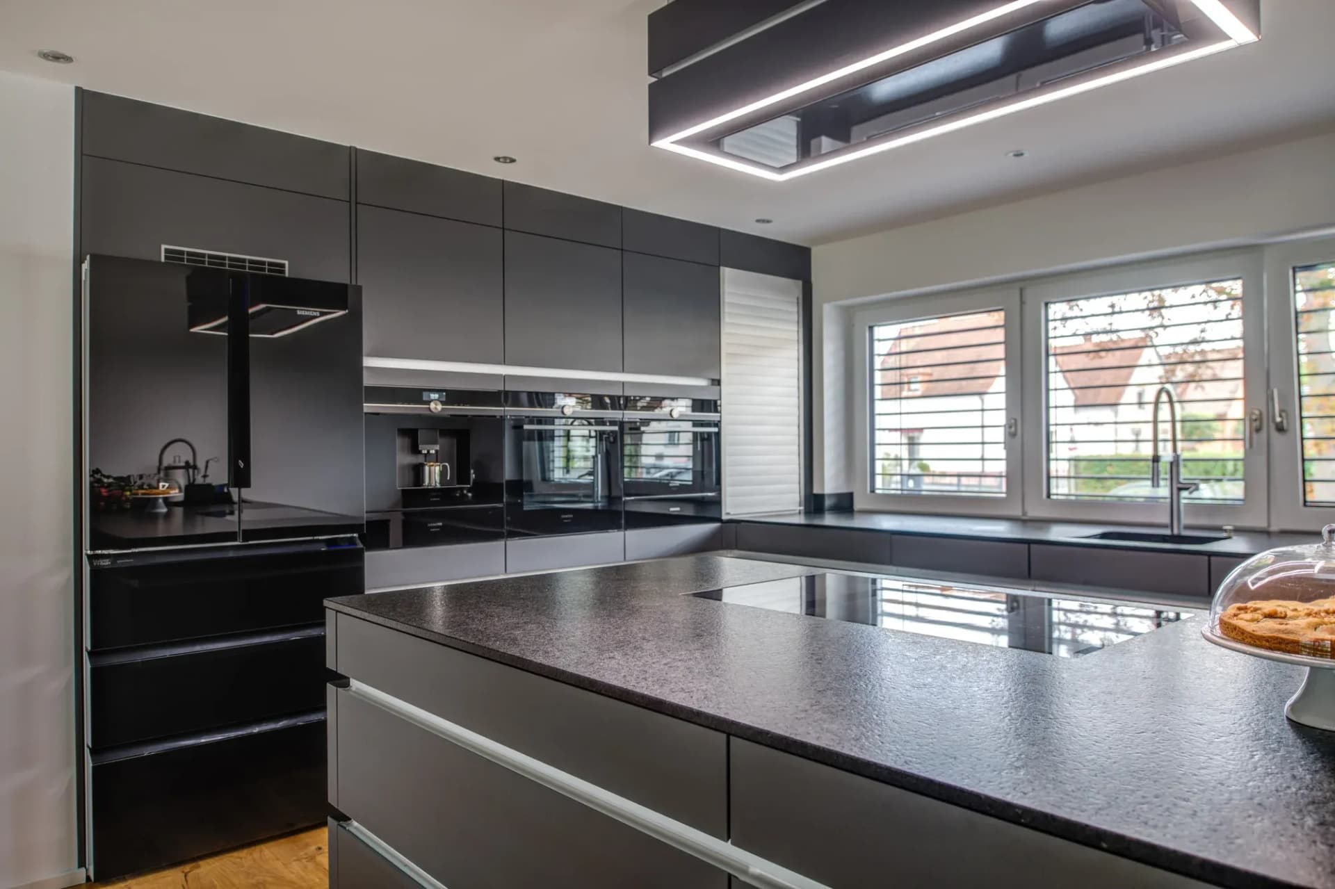 Blick über die schwarze Granit-Arbeitsplatte der Kochinsel auf die Gerätezeile mit Backöfen und Kaffeevollautomat, darüber eine beleuchtete Deckenhaube.