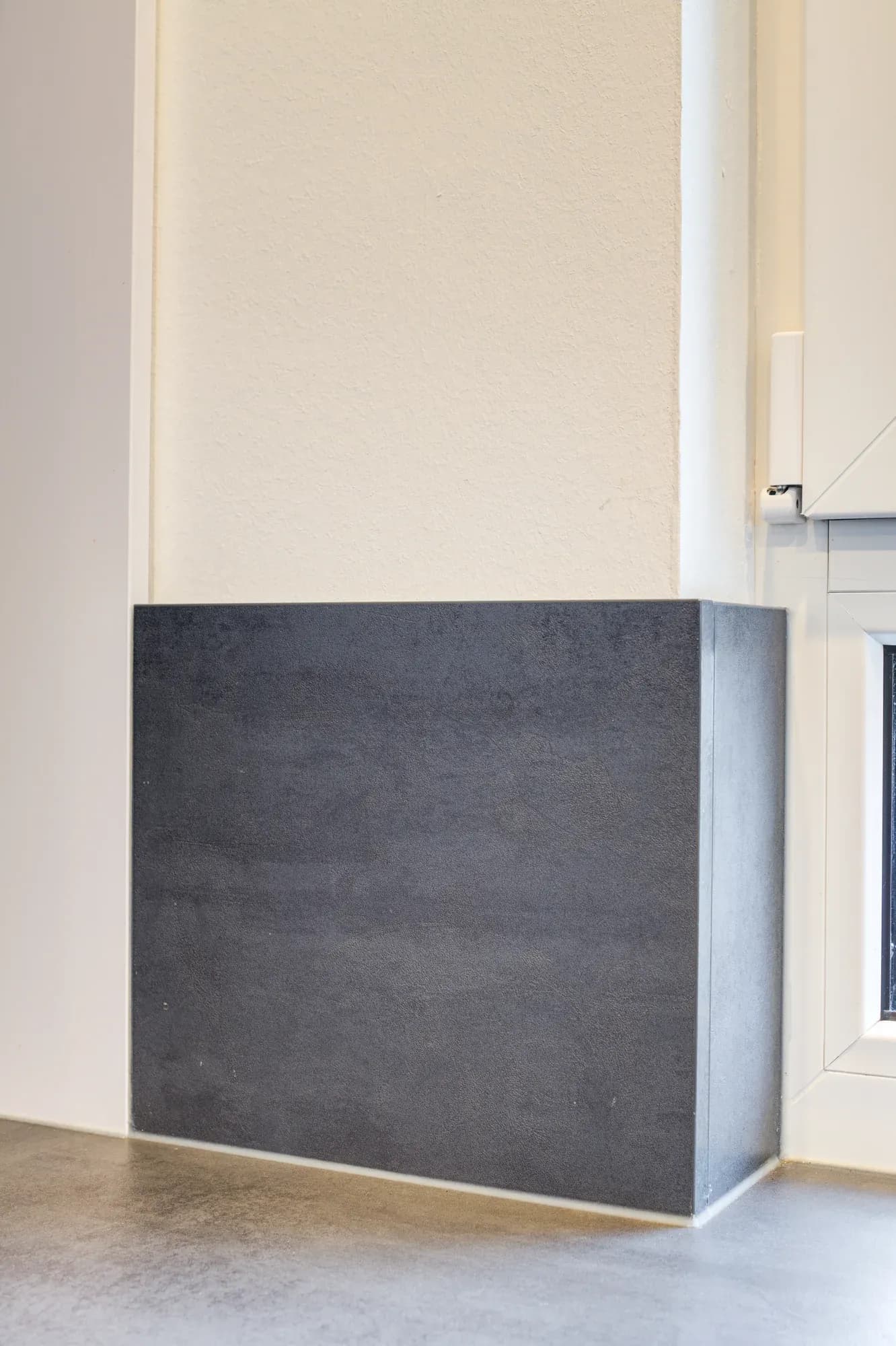 Niedriger, mit dunkler Keramik verkleideter Raumteiler-Block vor weißer Wand auf hellem Eichenboden.