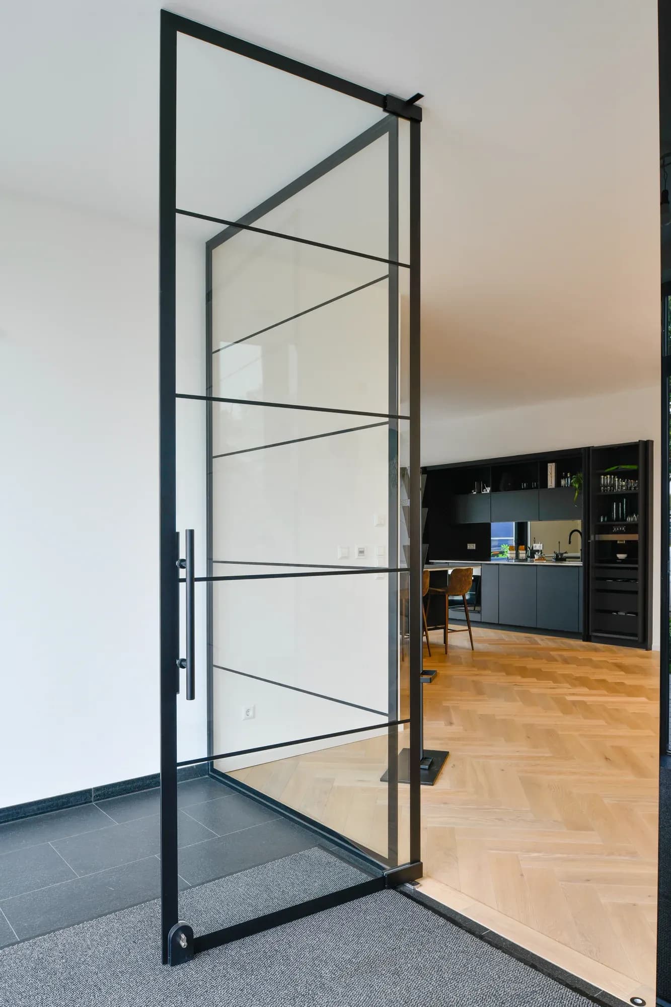 Loft-Tür aus schwarzem Stahlrahmen und Glas, die vom Eingang mit Schieferboden in die Büroräume mit Fischgrät-Parkett führt.