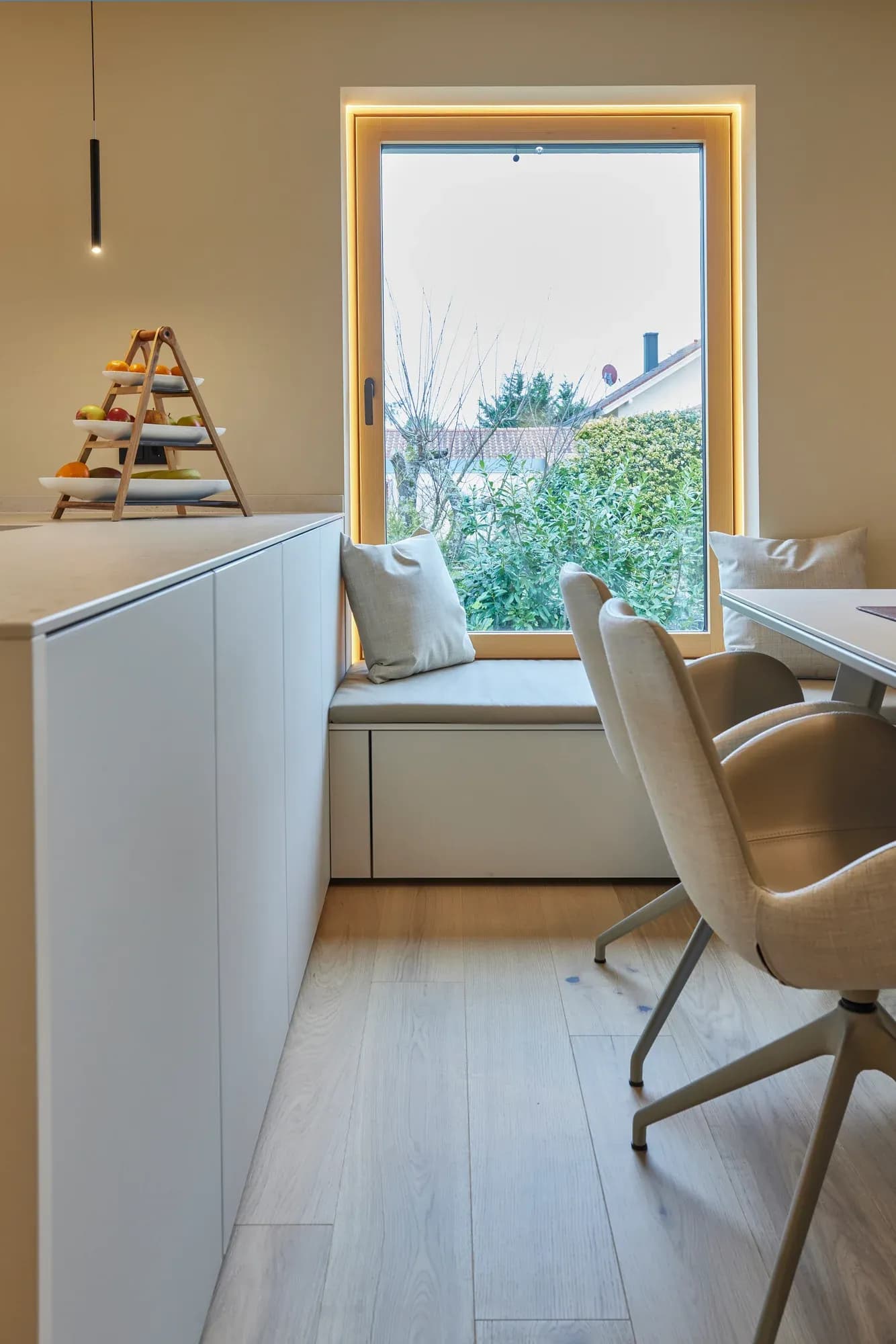 Gepolsterte Sitzbank in der Fensternische mit indirekt beleuchteter Laibung; davor ein heller Polsterstuhl am Esstisch, daneben eine weiße Sideboard-Zeile.