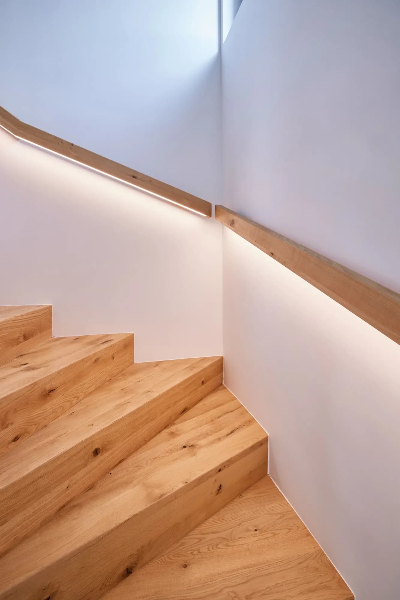 Massivholztreppe mit hinterleuchtetem Handlauf, der das Licht entlang der Wandecke führt.