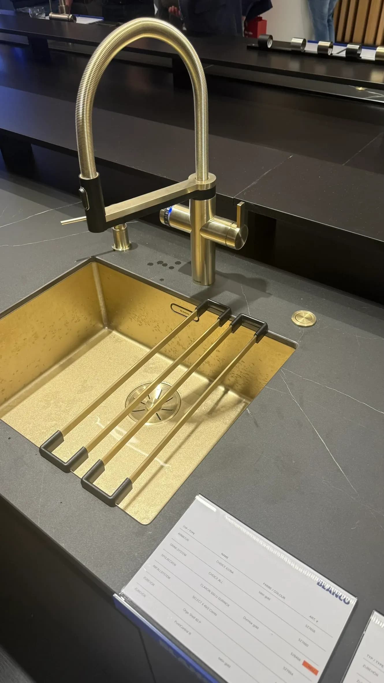 Bei Blanco auf Gut Böckel auf der Küchenmeile 2026: Spülbecken CLARON 500-U in DURINOX gold mit der Armatur CHOICE ICONA in satin gold, im Vordergrund die Produkttafel.