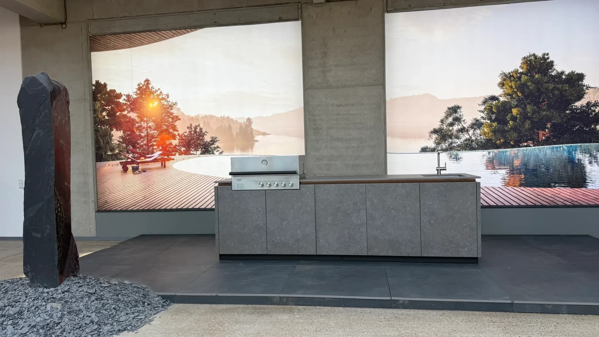 Bei BURN Outdoor Kitchen in Bissendorf auf der Küchenmeile 2026: Outdoorküchenzeile mit grauen Fronten, Edelstahlgrill und Spüle vor einer großformatigen Landschaftsfototapete.