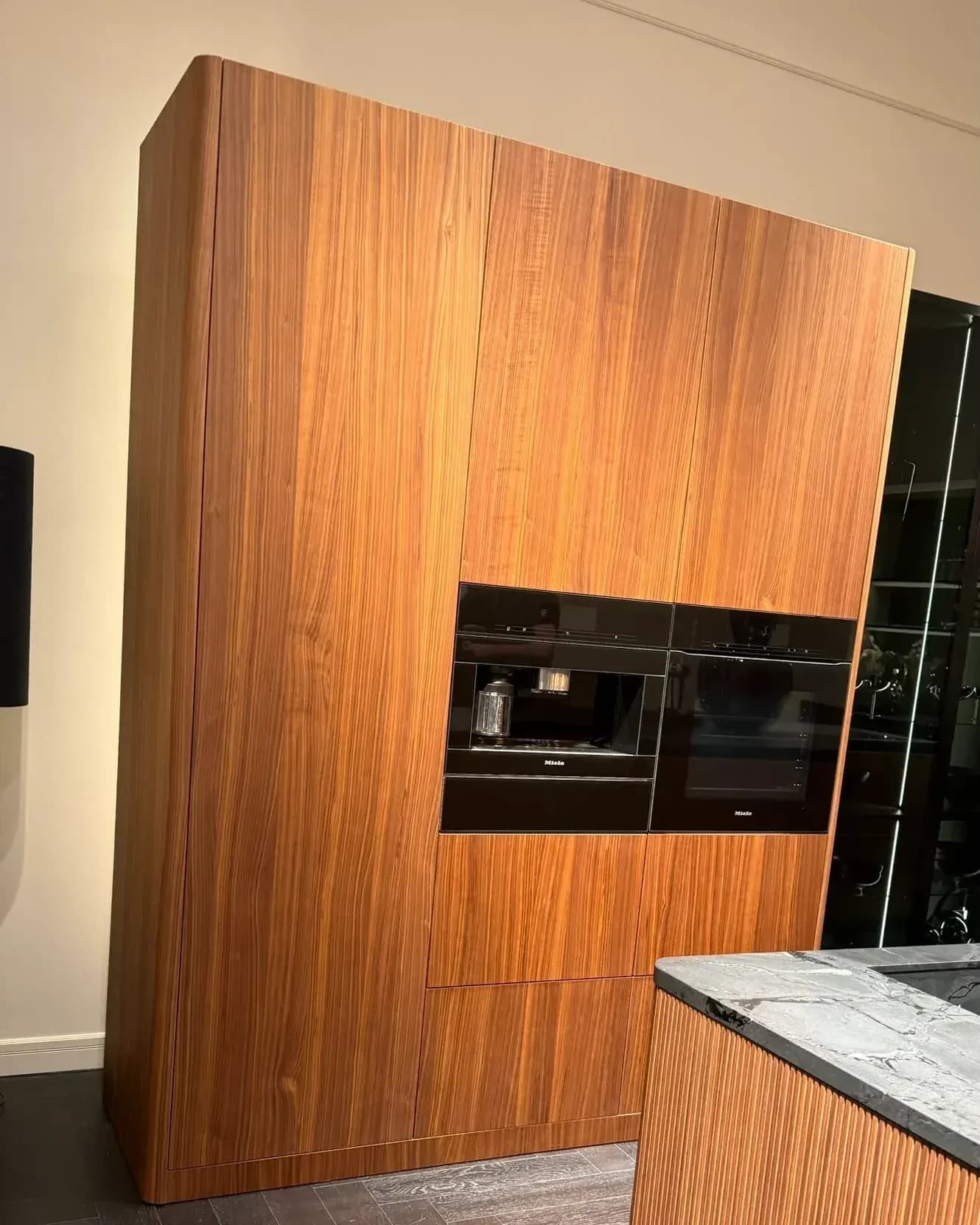 In der Hausausstellung von Häcker Küchen auf der Küchenmeile 2026: Hochschrankzeile in Nussbaum mit abgerundeter Seitenwange und zwei Einbaugeräten, im Vordergrund eine Insel mit gerillter Holzfront.