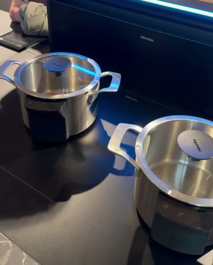 Messestand von Miele auf der Küchenmeile 2026: zwei Edelstahltöpfe auf einem schwarzen Kochfeld, dahinter ein hochgefahrener Dunstabzug mit blau leuchtender Kante.