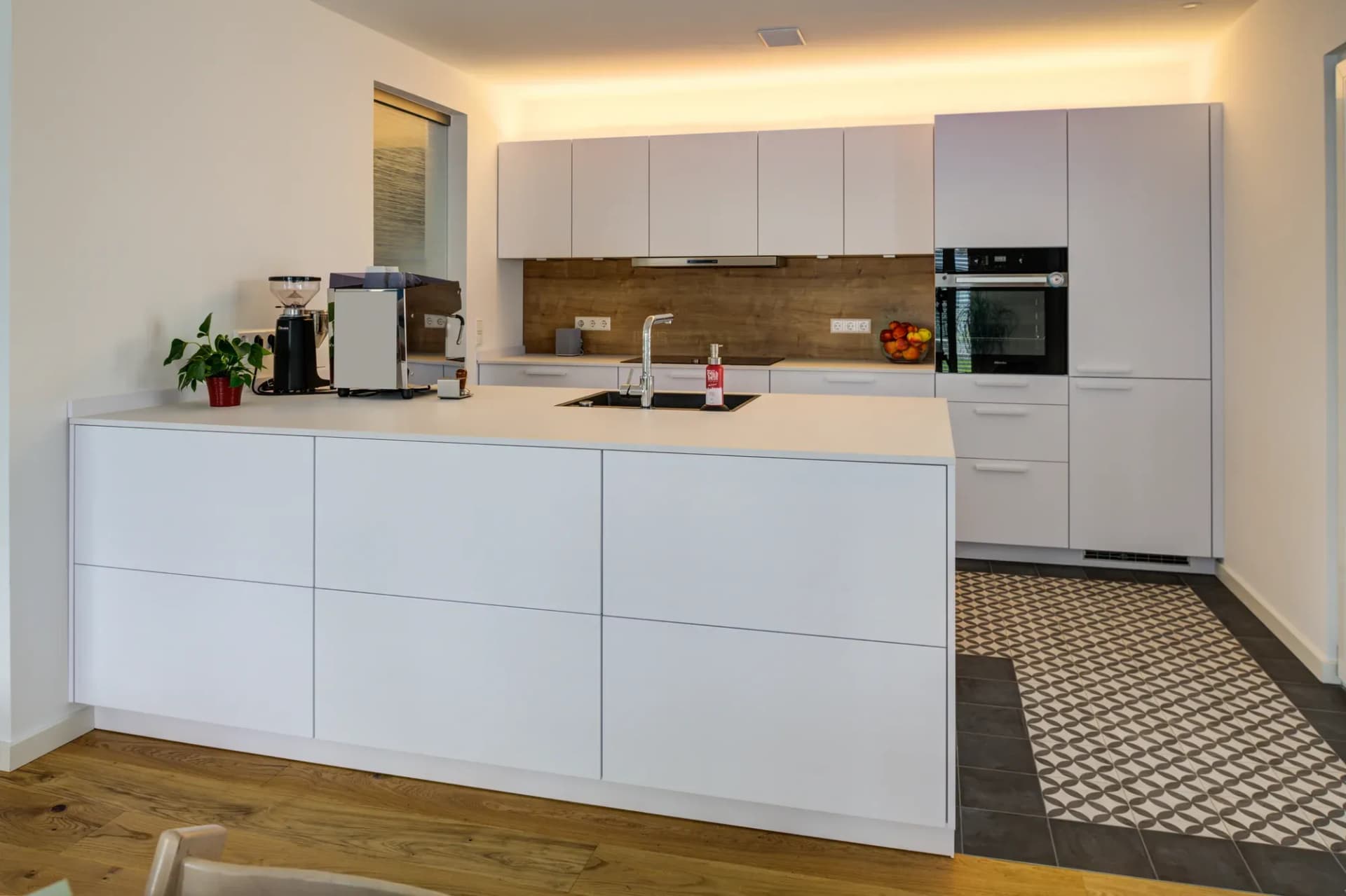 Offene Küche mit weißen Mattfronten, freistehender Insel mit heller Arbeitsplatte und schwarzer Granitspüle, Holzoptik-Rückwand und Miele-Einbaubackofen; im Übergang ein Boden aus gemusterten Zementfliesen.