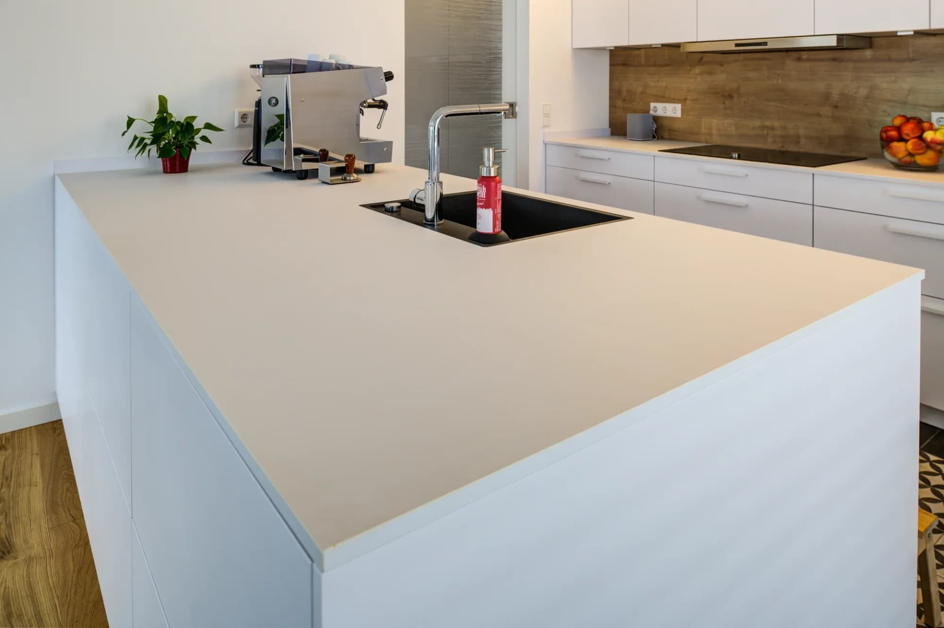 Detail der Kücheninsel: helle Arbeitsplatte mit eingelassener schwarzer Granitspüle und Chromarmatur, dahinter die Zeile mit Holzoptik-Rückwand.
