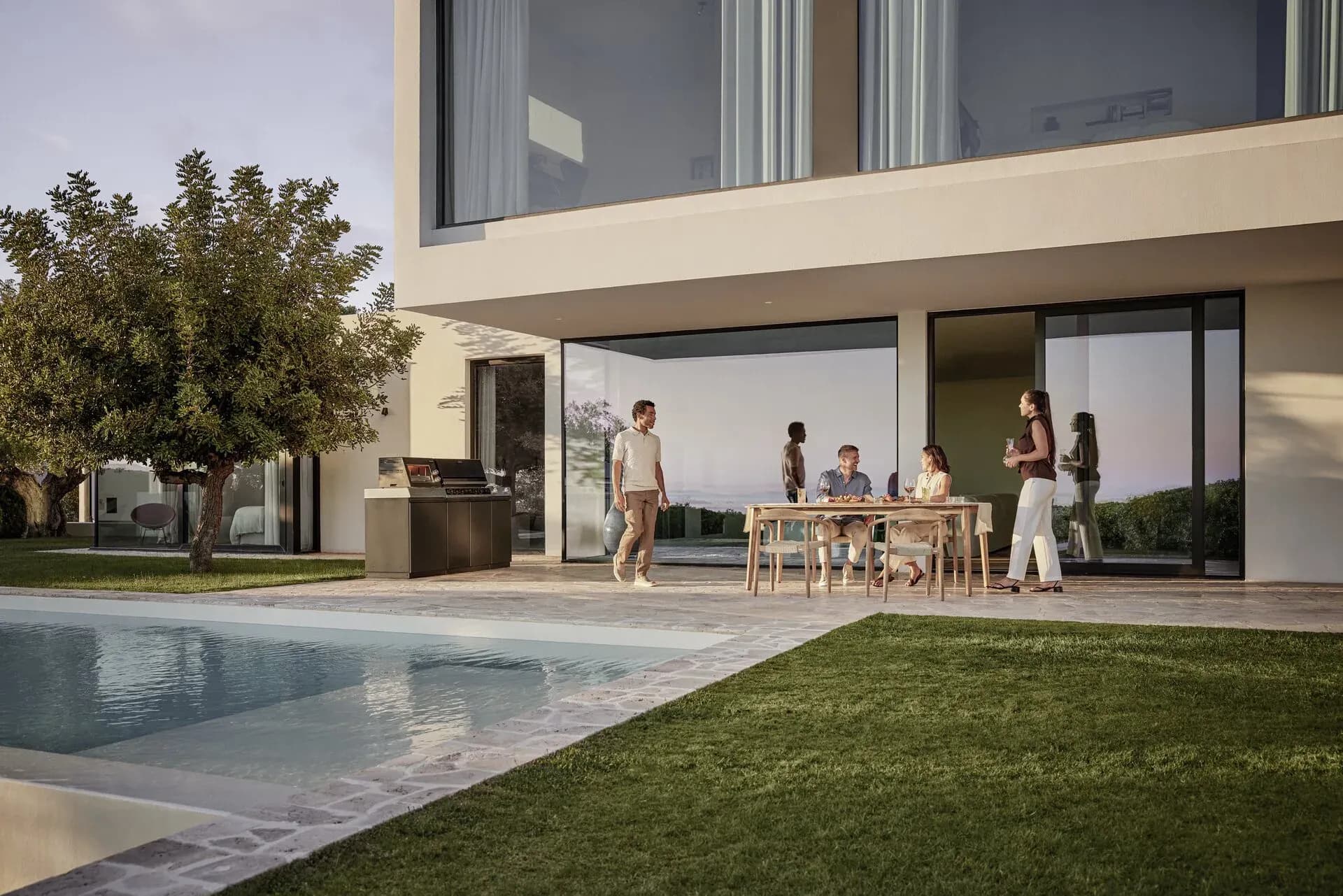 Freunde essen in der Abenddämmerung an einem Holztisch auf der Terrasse einer Villa, daneben eine Miele-Outdoor-Küche am Pool