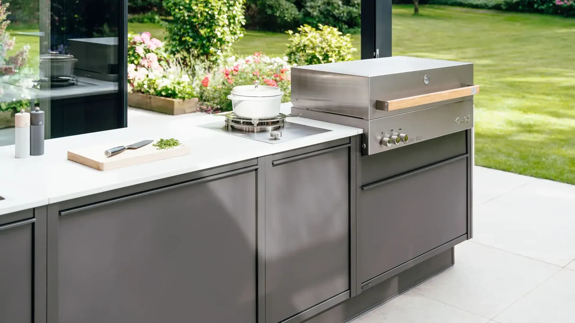 Anthrazitfarbene Outdoor-Küche mit Edelstahlgrill, Kochfeld und Schneidebrett auf einer hellen Terrasse vor dem Garten