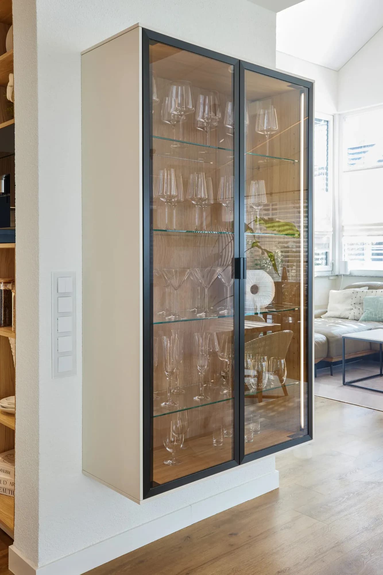 Schwarz gerahmte Glasvitrine mit eichefurniertem Innenraum und Glasböden, gefüllt mit Wein- und Sektgläsern, dahinter der lichte Wohnbereich mit Sofa.