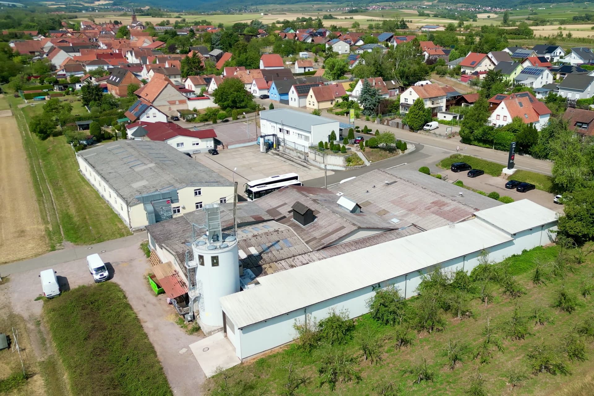 Drohnenaufnahme der Schreinerei in Schweighofen im Sommer: das Spänesilo an der weißen Werkhalle, dahinter das Dorf, davor Obstbaumreihen.
