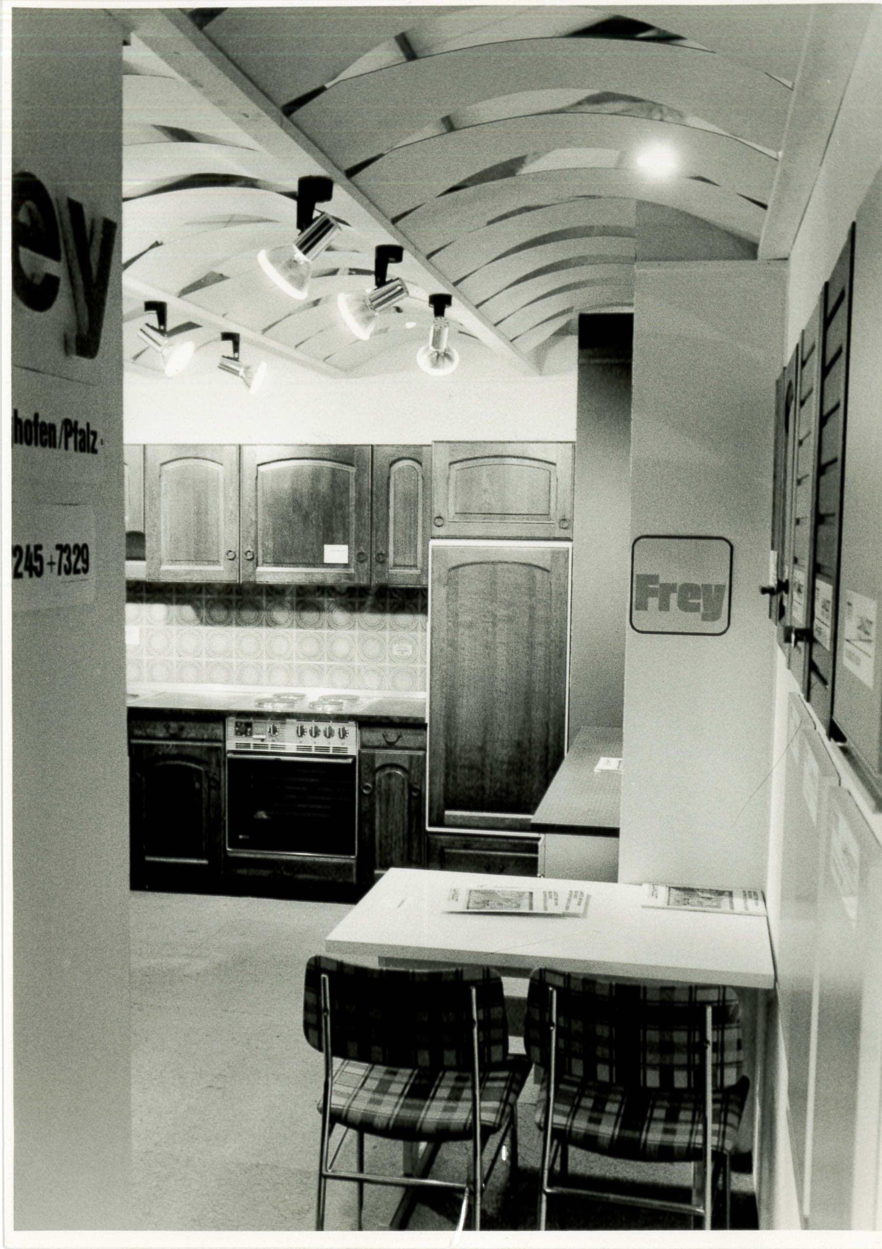 Die Schweighofener Küchenausstellung in Schwarz-Weiß: dunkle Eichenküche unter einer geschwungenen Lamellendecke, an der Säule ein Frey-Schild.
