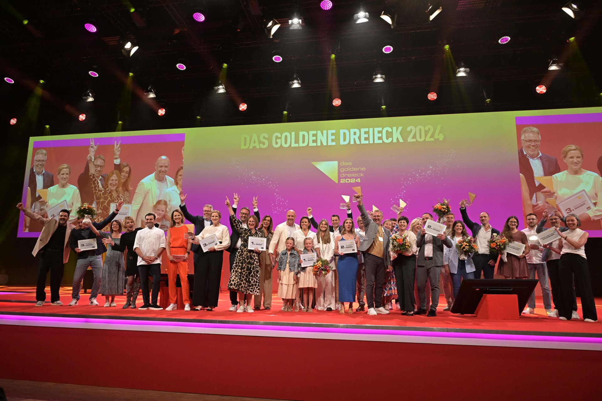 Preisverleihung „Das Goldene Dreieck 2024" auf der Bühne: die Gewinner-Teams mit Urkunden und Pokalen vor der Bühnenleinwand