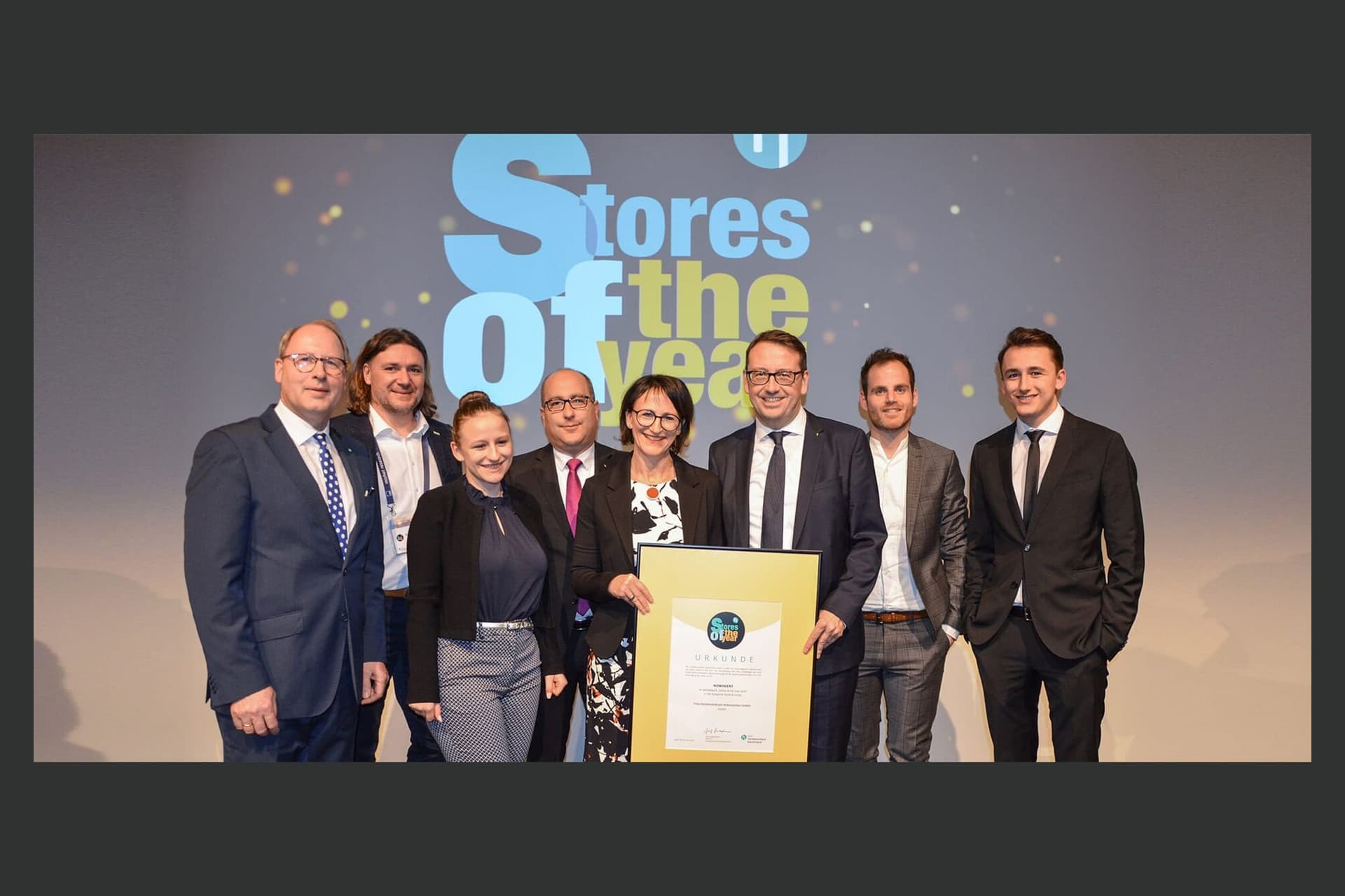 Bühnenfoto der Verleihung „Stores of the Year" 2019 in Berlin: acht Personen vor der Leinwand, vier halten die gerahmte Urkunde.