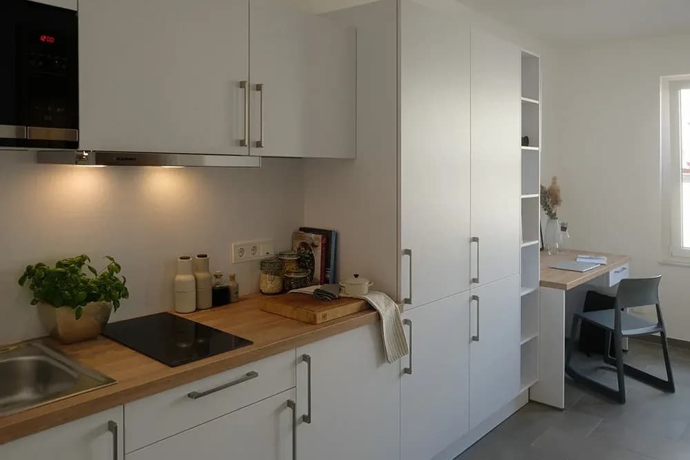 Studierenden-Apartment: weiße Küchenzeile mit Holz-Arbeitsplatte, Hochschränken mit Regalnische und bündig anschließendem, integriertem Schreibtisch.