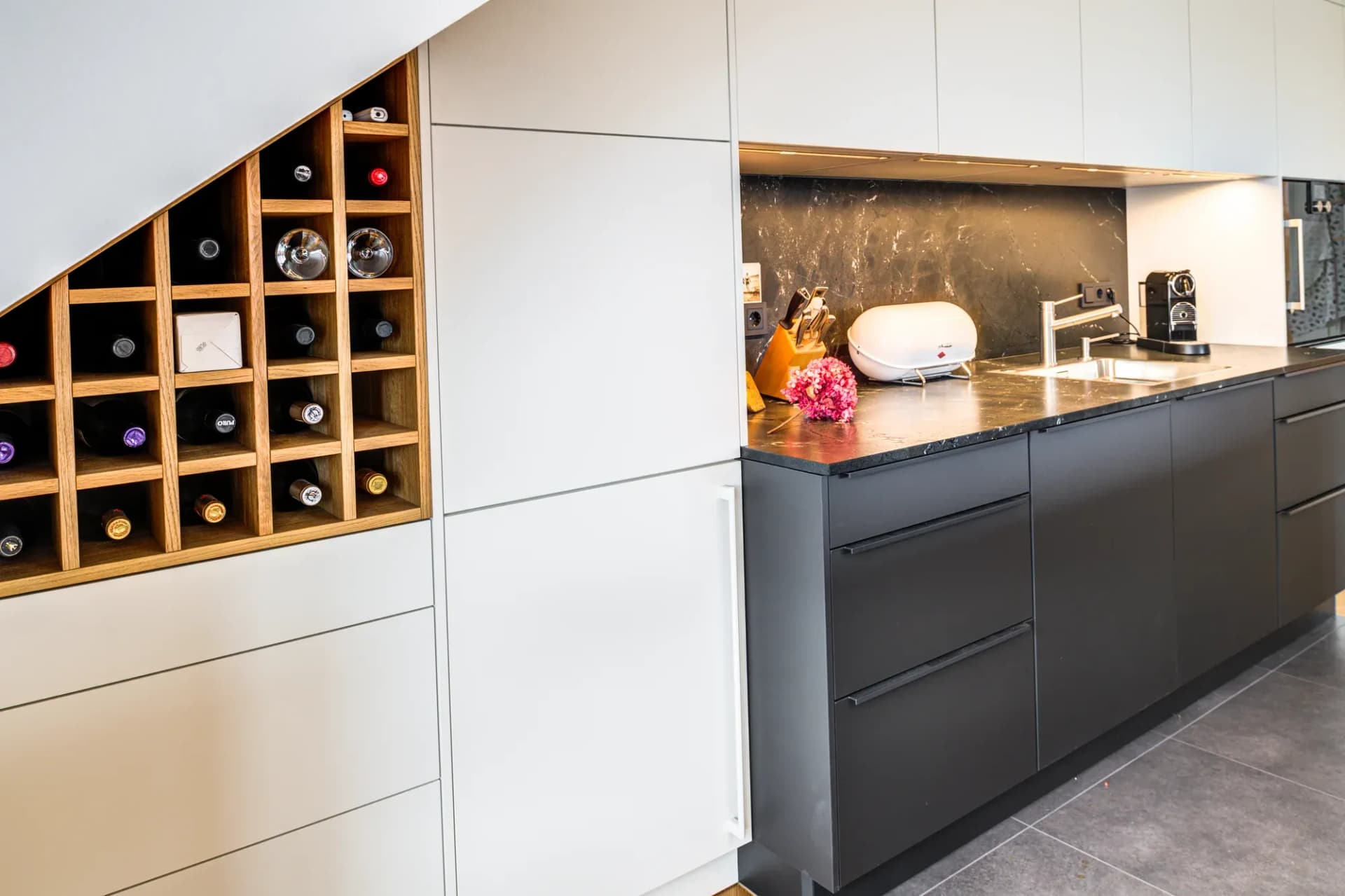 In die Dachschräge eingepasstes Weinregal aus Eiche neben der grifflosen hellen Schrankwand, daneben die anthrazitfarbene Arbeitszeile mit schwarzem Naturstein.