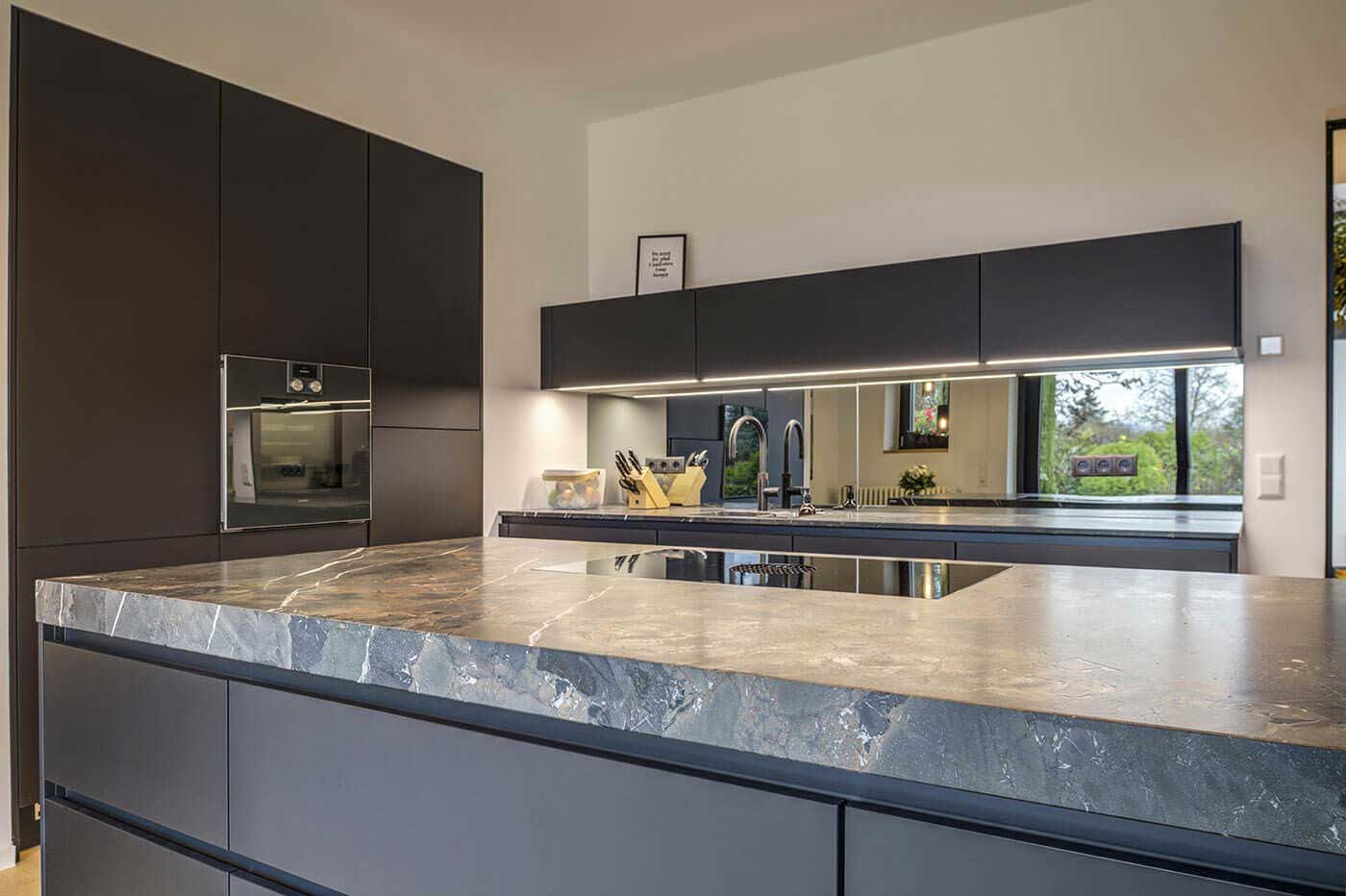 Moderne dunkle Küche mit Arbeitsplatte in Stein-Optik, Kochfeld mit integriertem Dampfabzug und Unterschrankbeleuchtung