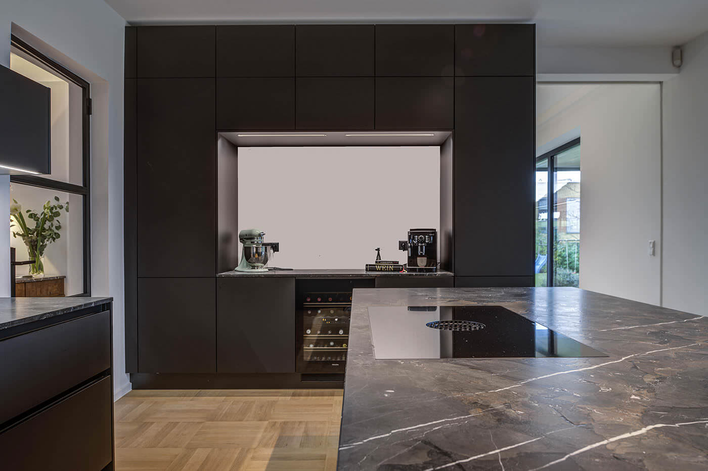 Moderne dunkle Küche mit Arbeitsplatte in Stein-Optik, Kochfeld mit integriertem Dampfabzug, stylischen Geräten, Weinkühler und Durchreiche