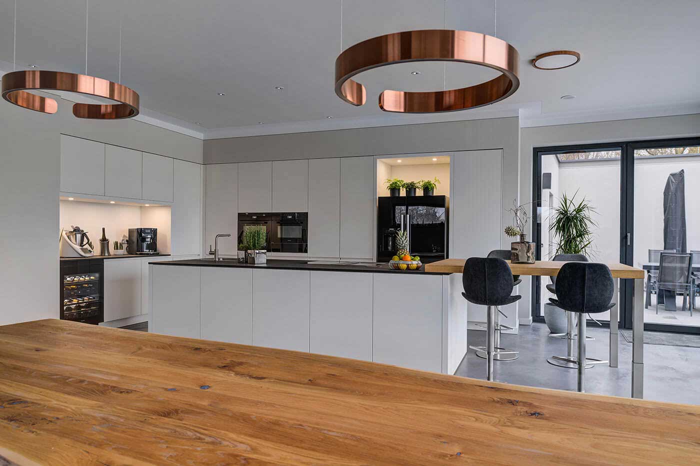 Moderne weiße Küche mit dunkler Arbeitsplatte, stylischen Elektrogeräten, Weinkühler, Holztresen und Esszimmertisch aus Massivholz
